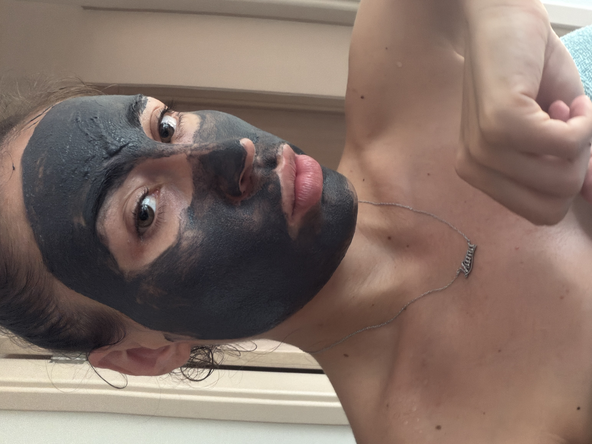 A woman is applying a charcoal face mask in this close-up, bust-level portrait. Her eyes are open and looking directly at the camera. The charcoal mask covers most of her face, with her lips and parts of her eyes visible. She is wearing a silver necklace with a nameplate that reads "Llana". The background appears to be a room with beige walls and vertical paneling, possibly a bathroom or bedroom. The lighting is soft and natural, suggesting it is daytime. The woman's skin tone is fair, and her hair is pulled back. Her left shoulder and upper chest are visible, as is her right hand, which is held up near her chin. The overall impression is one of self-care and relaxation. The image was taken in the city of Tersefanou, Cyprus.
