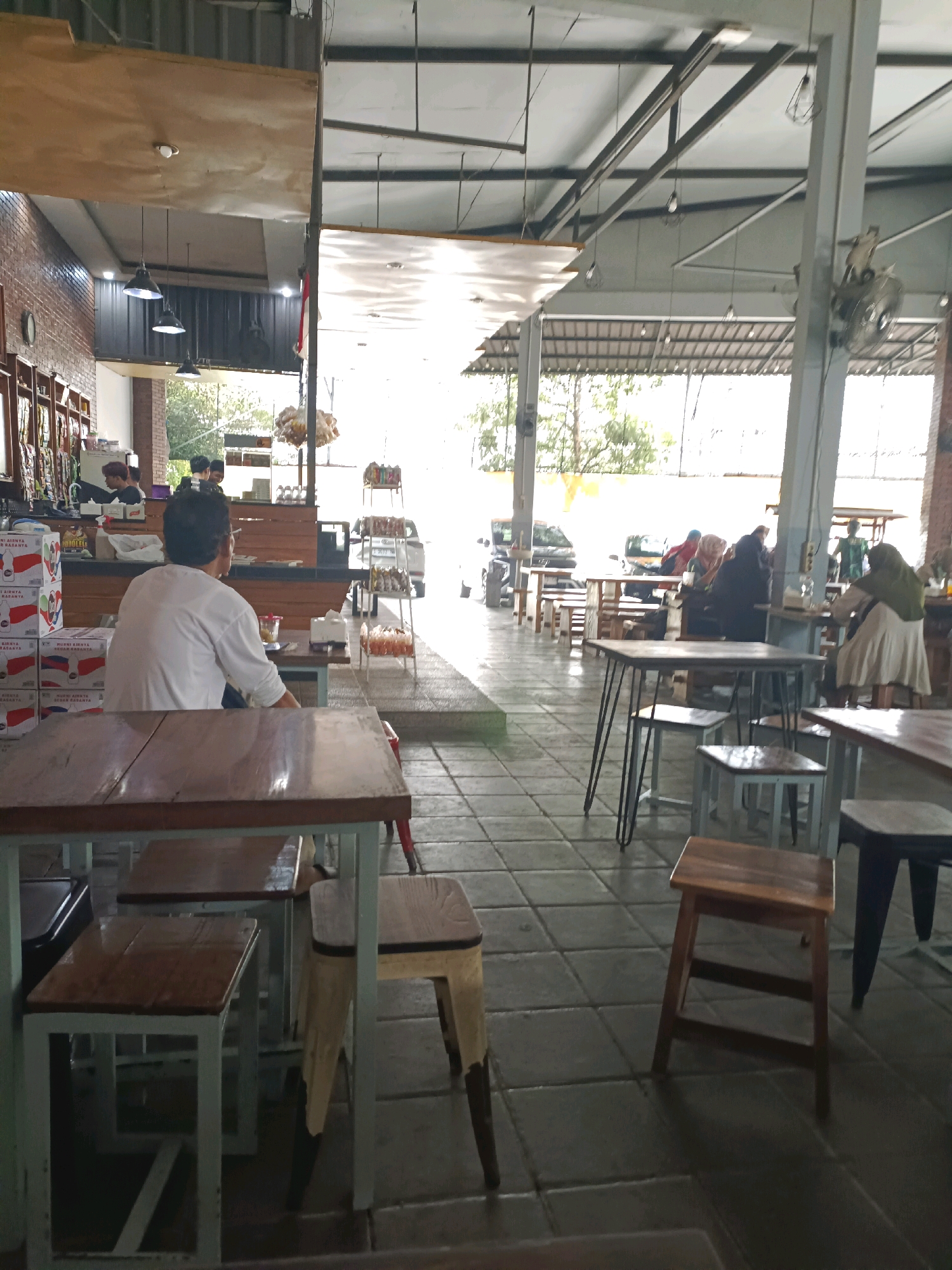The image depicts the interior of a busy food court or cafe in Made, Indonesia. The setting appears to be open-air with a covered roof supported by industrial metal beams. Natural light streams in from the sides, illuminating the tiled floor and various seating areas.
In the foreground, a man in a white shirt is seated at a table, facing away from the camera. He is the central figure in this part of the frame. Several wooden tables and chairs, some with metal frames, are visible throughout the space, indicating a dining establishment.
Further back, other patrons are seen engaged in activities typical of a cafe. A woman in a green hijab and light-colored dress is standing near a table. Other individuals, some wearing headscarves, are seated at various tables.
On the left side of the image, there are shelves stocked with items, possibly merchandise or food products, suggesting a retail or complementary service within the establishment. Boxes with text and graphics are stacked near the counter area.
The overall atmosphere is casual and active, with a mix of people enjoying food and drinks in a semi-outdoor environment. The architecture combines brick elements with exposed metalwork, giving it a modern industrial feel. The presence of trees and foliage visible through the open sides indicates proximity to an outdoor area or a well-lit interior space.