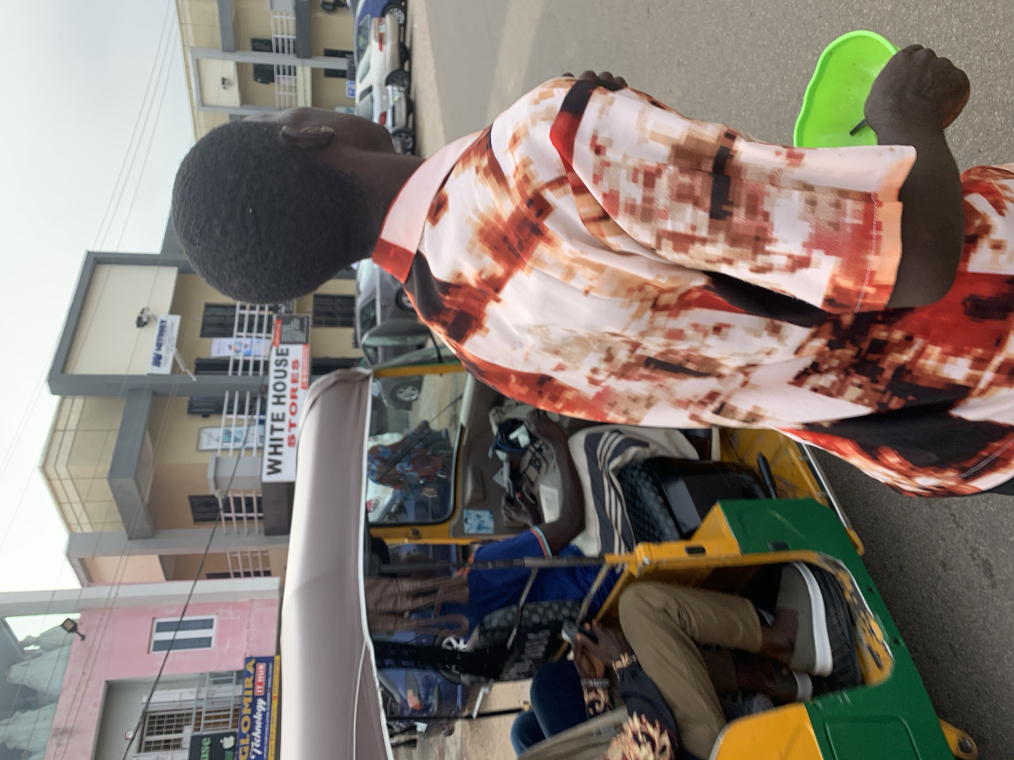 In Jos, Nigeria, a man with dark skin and short black hair walks away from the camera, his right arm bent and holding a bright green plastic container. He wears a colorful short-sleeved shirt with a pattern of brown, red, and white splotches. Behind him, a yellow and green auto-rickshaw is parked. Inside the auto-rickshaw, a person wearing a blue shirt and striped trousers drives, and another person in the passenger seat wears khaki pants and a patterned shirt. The auto-rickshaw is parked on a paved street with several cars visible in the background. Buildings with various colored facades, including pink, beige, and gray, line the street, with signs indicating businesses such as "GLOMIRA Technology IT HUB" and "WHITE HOUSE STORES". The sky is overcast.