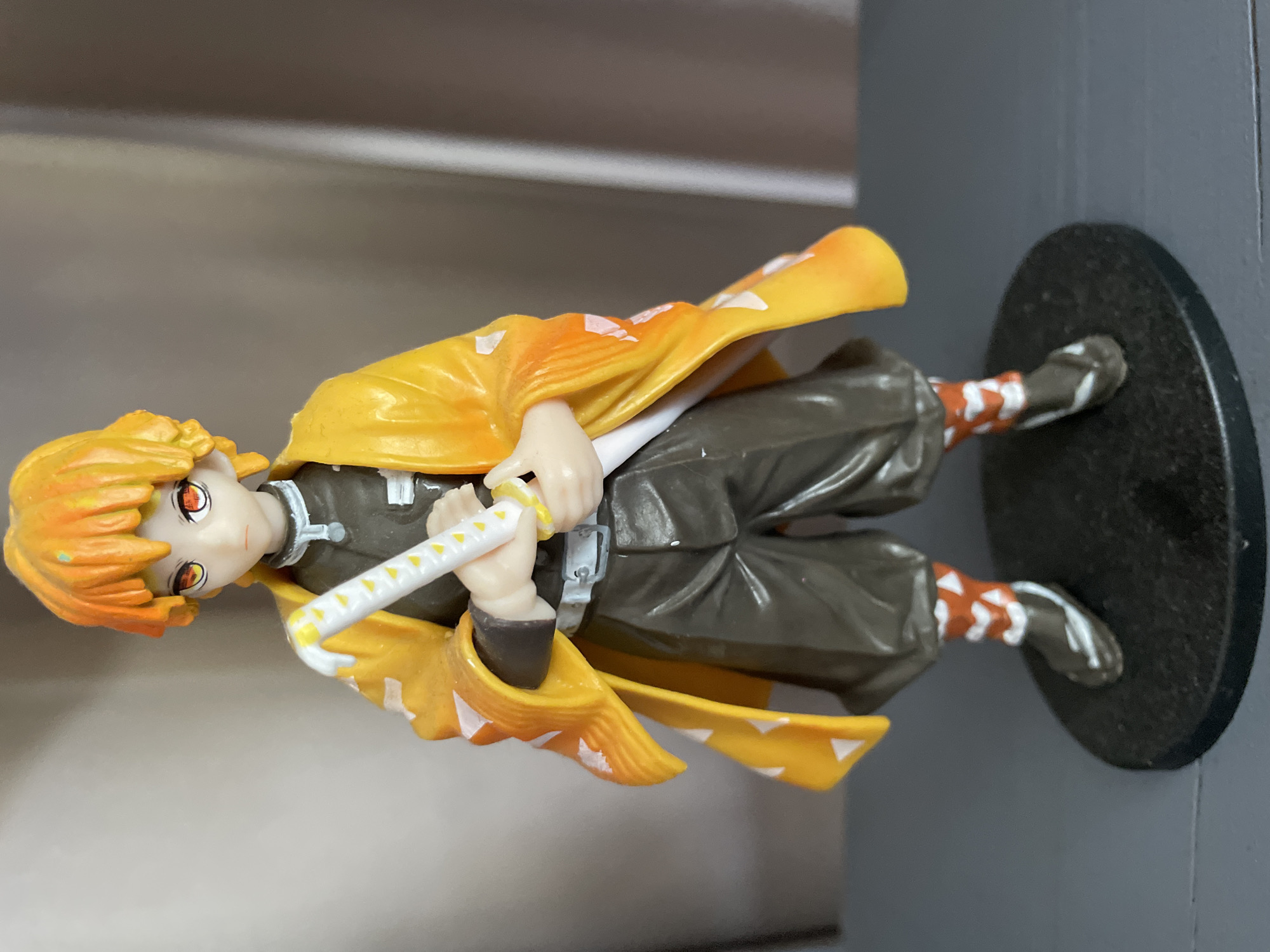 A full shot captures a detailed figurine of a young male anime character, likely from the series "Demon Slayer," standing on a black circular base. The character has spiky orange hair, bright orange eyes with red irises, and is wearing a dark brown uniform. Over the uniform, he sports a vibrant yellow haori (a traditional Japanese jacket) adorned with white triangular patterns. He is holding a white katana with yellow accents in both hands, positioned in front of him. His legs are clad in dark brown pants and orange and white striped socks, with dark shoes. The background consists of a softly blurred curtain, suggesting an indoor setting. The lighting appears to be from the front, casting subtle shadows. The image does not provide any information or context related to Sochi, Russia.