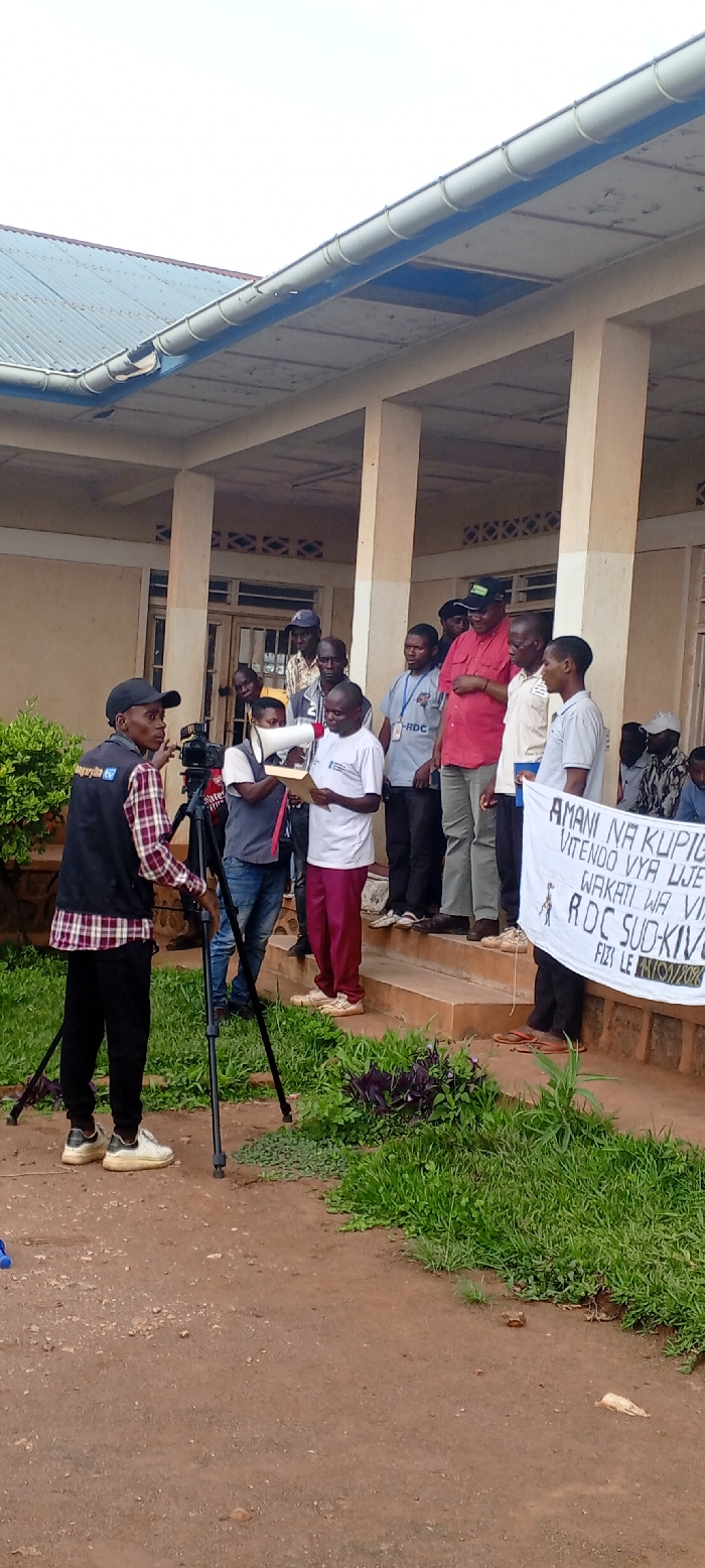 An outdoor gathering is documented in Kundu 1, Democratic Republic of the Congo, at what appears to be a public building. The building features light-colored walls, multiple support pillars, and a covered porch area with a corrugated metal roof and white gutters.
In the foreground, a male individual, dressed in a plaid shirt, black "Suguriyata TV" vest, black pants, and white sneakers, operates a video camera mounted on a black tripod. He is positioned on a reddish-brown dirt ground with patches of green vegetation.
On a set of concrete steps leading to the building's entrance, a group of approximately 15-20 adult males is assembled. A man in a white t-shirt and burgundy pants holds a white megaphone in his right hand and a brown document or folder in his left, facing leftward as if addressing the camera. To his right, another man in a red polo shirt and dark cap stands. Further to the right, two individuals hold a white fabric banner with black text, partially visible, which includes phrases such as "AMANI NA KUPIGA," "VITENDO VYA UJE," "WAKATI WA VI," "RDC SUD-KIVU," and "FIZI LE 24/01/202X." Other men are clustered behind and around these central figures, some wearing identification badges. The sky is overcast.