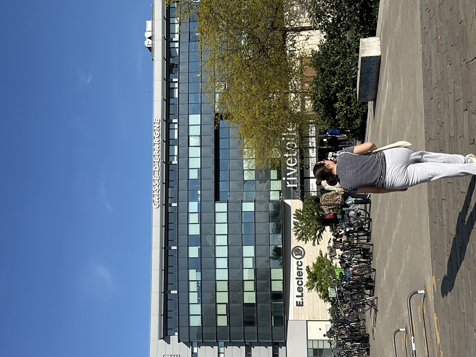 A horizontal, eye-level shot captures a tall, modern building in Strasbourg, France, against a clear blue sky. The building, identified as the "Caisse d'Epargne" and the "E.Leclerc Rivetoile," is a multi-story structure with a facade of large, reflective windows in a grid pattern. In front of the building, there's a parking area filled with parked bicycles. A woman dressed in a striped shirt and gray pants is standing on a raised, paved platform in the foreground. The platform is made of gray stone or concrete blocks. There is a tree on top of the parking area. The building has the sign "rivetoile" and then, underneath, in smaller print, is the text: "PHORA/COUSS." The "E.Leclerc" sign is circular, with the letter "L" in white on a blue background.