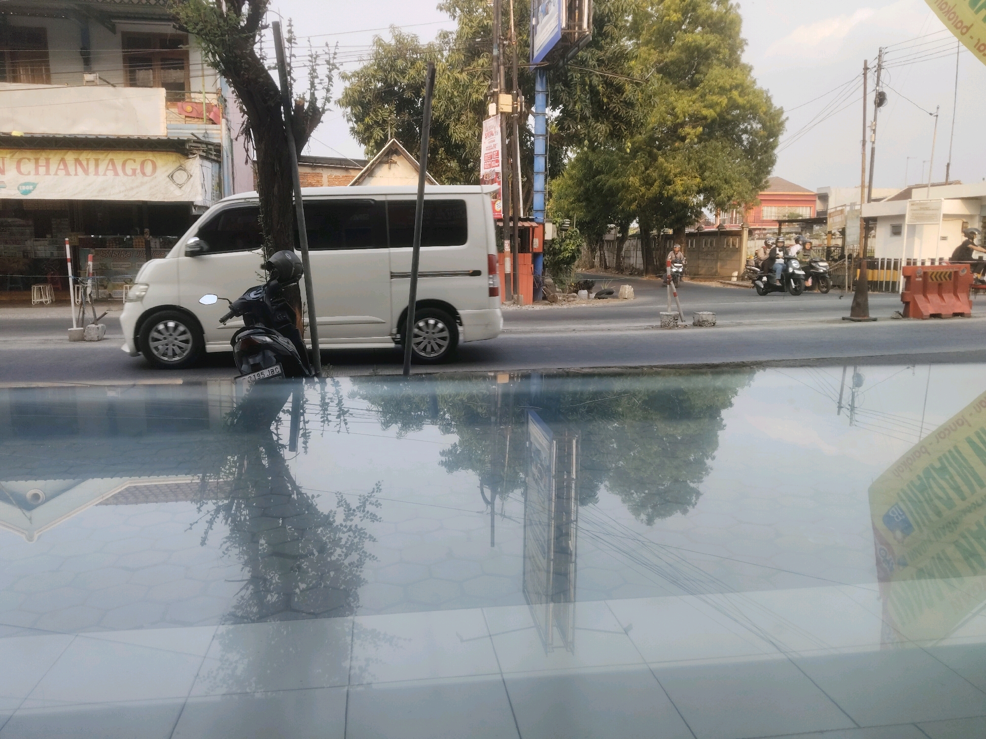The image captures a street scene in Sragen, Indonesia, reflected on a wet, glassy surface. In the foreground, the reflection shows a tree, overhead power lines, and a tall billboard with Indonesian text. The actual scene, visible above the reflection, features a white van parked on the side of a road. A motorcycle with a rider wearing a helmet is parked next to the van. Further down the road, several people are riding on motorcycles. Buildings line the street, and lush green trees are abundant. The sky is overcast. The overall impression is of a typical day in an Indonesian town, with traffic and daily life unfolding along the roadside.