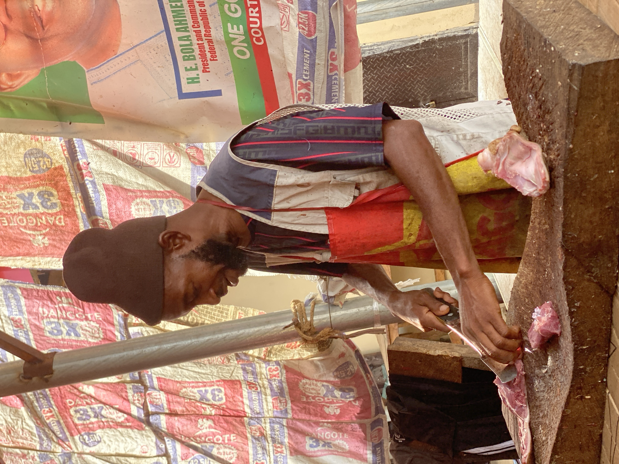 The image shows a man, likely a butcher or meat vendor, engrossed in cutting red meat on a sturdy wooden chopping block. He is wearing an apron over a patterned short-sleeved shirt and a dark cap, and uses a knife to process the meat. The scene takes place under a makeshift shelter, possibly a market stall, in Kafanchan B, Nigeria.
The walls and roofing of the stall are constructed from numerous repurposed polypropylene sacks. Many of these sacks are clearly identifiable with the branding "DANGOTE 3X CEMENT 42.5N," indicating they are cement bags. Another large sack, serving as part of the canopy, displays political text including "H. E. BOLA AHMED," "President and Commander in Chief," and "Federal Republic of," which likely refers to Nigeria's current president, Bola Ahmed Tinubu. Other visible text includes "ONE GO" and "COURT."
The bright, natural lighting suggests it is daytime. A metal pole, part of the structural support, traverses the mid-ground. The overall impression is of a focused individual engaged in a daily trade within an active, informal market environment.