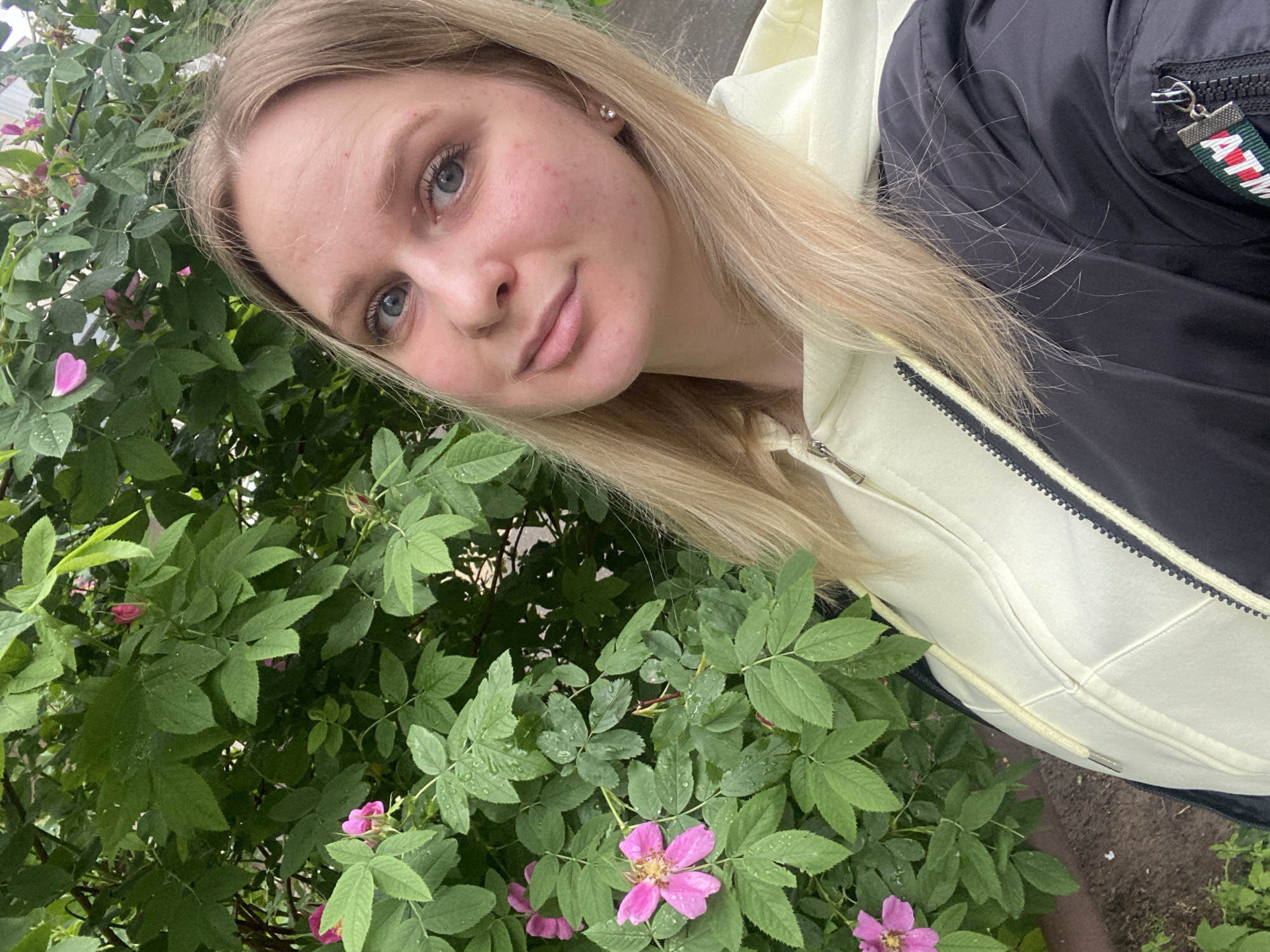 An outdoor, eye-level selfie-style photograph depicts a young adult female positioned in front of a dense, green bush with pink flowers in the city of Unknown, Unknown. The subject, a female with long blonde hair and blue eyes, faces the camera with a slight smile. Her fair complexion shows minor facial blemishes on her cheeks and forehead. She wears a light yellow zippered hoodie under a black bomber-style jacket. The left sleeve of the black jacket features a utility pocket with a zipper and a fabric pull tab colored green, white, and red, partially displaying "ATM" in white text. A small, clear or silver stud earring is visible in her right ear. The background consists of copious green foliage with many leaves bearing visible water droplets. Several five-petaled pink flowers are interspersed throughout the leaves, notably one prominent bloom with a yellow/brown center on the mid-left side. A dark patch of earth is partially visible in the lower-left corner beneath the bush. The lighting is soft and even, suggesting an overcast day or post-rain conditions.