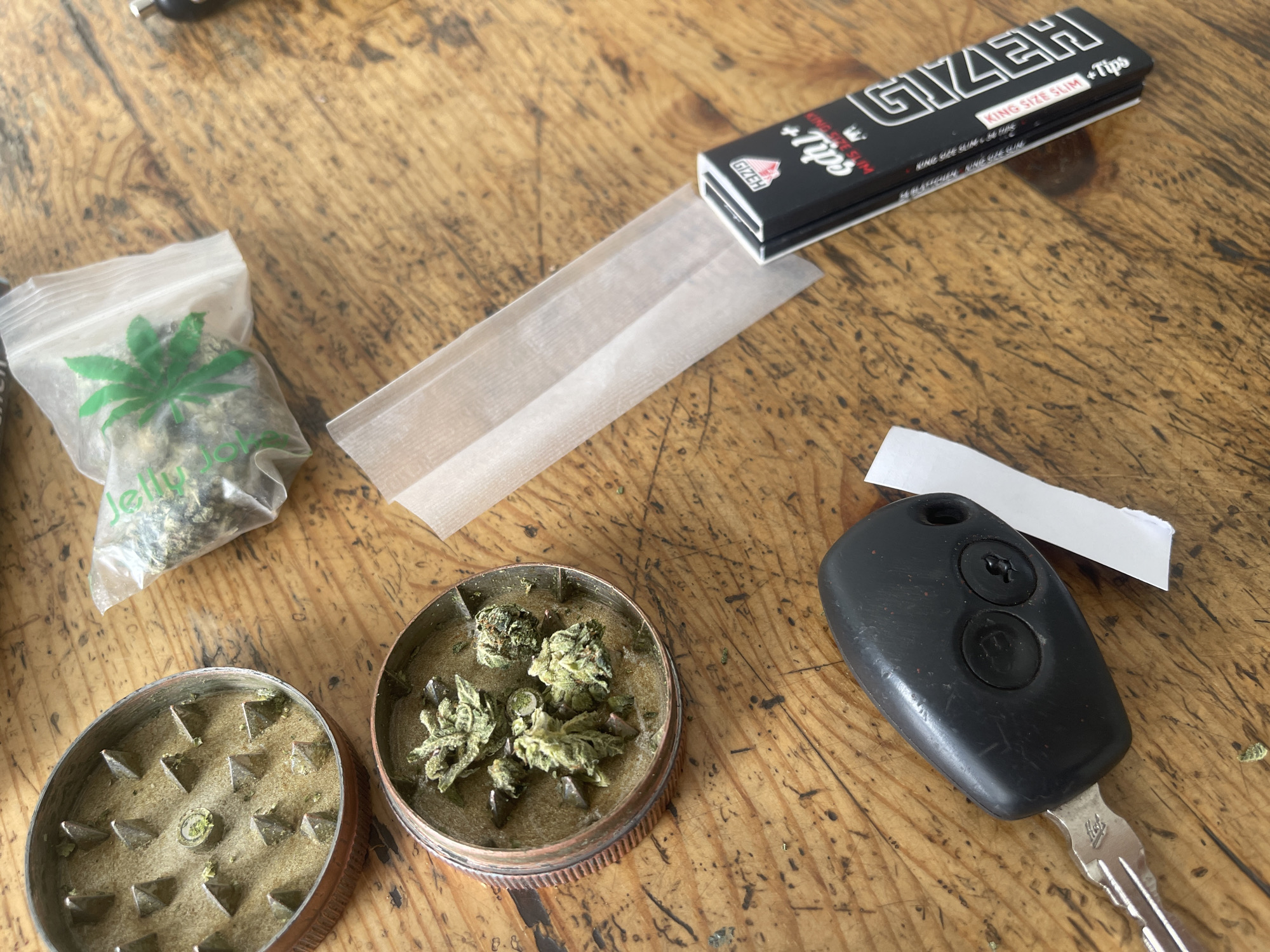 The image shows a wooden surface with several items related to cannabis use. In the upper left corner is a clear plastic bag containing cannabis flower, with a green cannabis leaf printed on it. To the right of the bag, there's a partially opened pack of Gizeh king-size slim rolling papers. Below the rolling papers, there is a metal grinder with one section containing cannabis flower and the other open, showing the grinding teeth. To the right of the grinder is a black car key. A small piece of white paper is next to the car key. The image suggests the preparation of a cannabis joint.