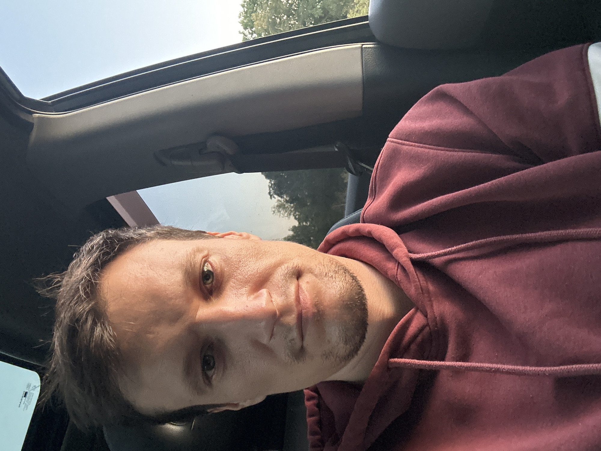 A close-up, selfie-style photograph captures a light-skinned man in his late 20s or early 30s sitting in the passenger seat of a vehicle. He is looking directly at the camera with a neutral expression. The man has short, brown hair that is slightly messy, a light stubble on his chin, and a light beard. He is wearing a dark red hooded sweatshirt with a drawstring. The interior of the car is visible, including a black seat, a grey pillar, and a glimpse of the sky through the windshield. Outside the car, through the passenger window, green trees and a clear blue sky are visible. The image appears to have been taken during daylight hours. The provided context states the location is Makiv, Ukraine.