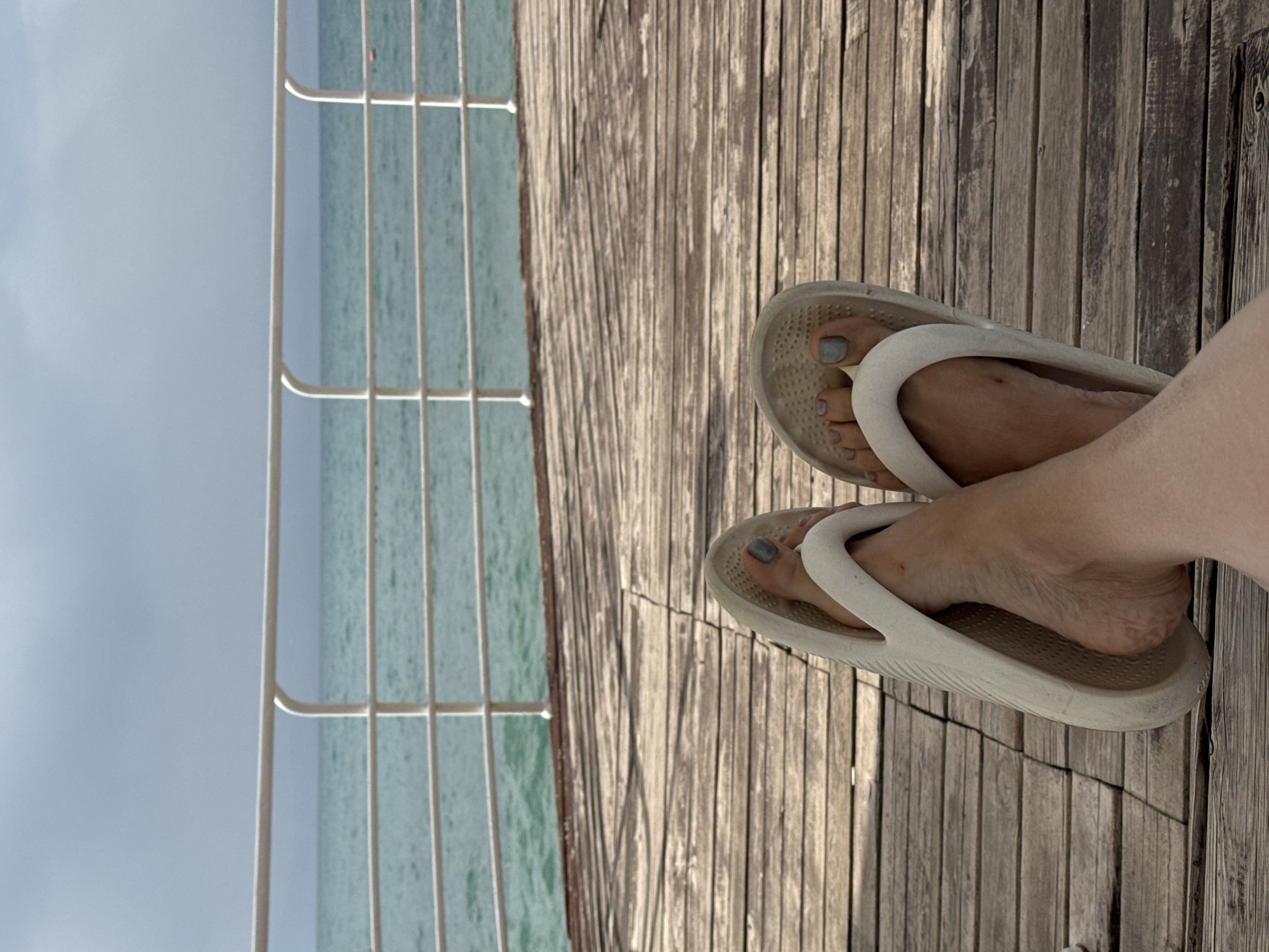 The image shows a close-up view of a person's feet resting on a wooden deck. The feet are adorned with grey nail polish and are encased in white flip-flops. In the background, a white railing separates the deck from a body of turquoise water, suggesting a location near a lake or the sea. The sky is overcast with grey clouds. The wooden planks of the deck are visible in the foreground and midground, oriented horizontally. The overall impression is one of relaxation in a waterside setting. This scene could plausibly be located in Sary-Oy, Kyrgyzstan, a resort area on the shores of Issyk-Kul Lake.