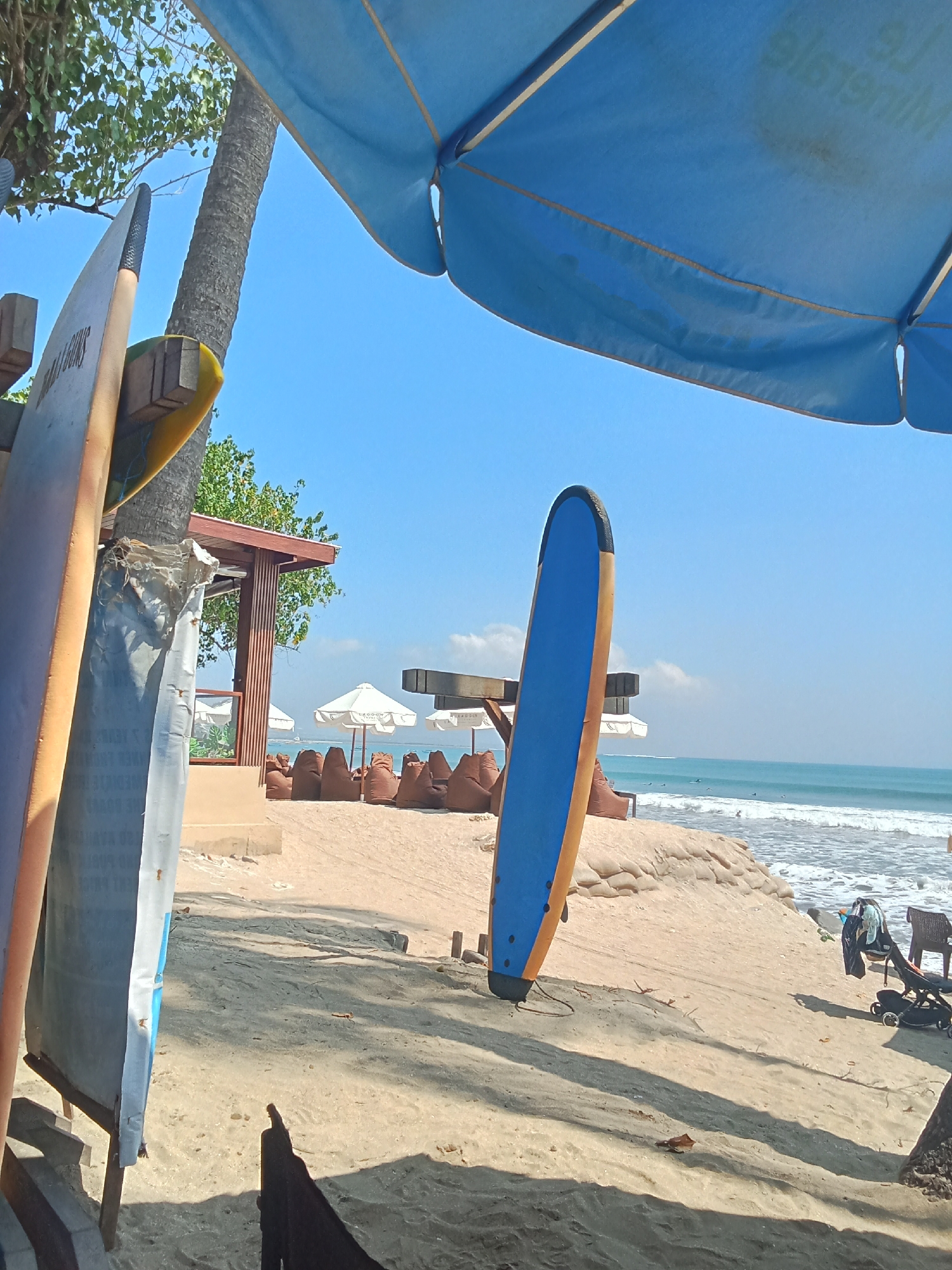 The image depicts a beach scene under a bright blue sky. In the foreground, two surfboards are propped upright, one on the left and a blue and orange one on the right. A large blue umbrella shades part of the scene from above. Behind the surfboards, a palm tree trunk is visible, and further back, a wooden structure with a sloped roof and vertical wooden slats stands near the beach. Several white umbrellas are set up on the sand, surrounding rows of brown beanbag chairs arranged facing the ocean. The ocean has moderate waves and several surfers can be seen in the water. The sand is light brown, and there are some sandbags forming a barrier on a slight incline leading down to the water. A stroller and a chair are also visible on the sand in the middle ground. The scene appears to be a relaxed, recreational area, possibly a beachside cafe or surf rental spot.