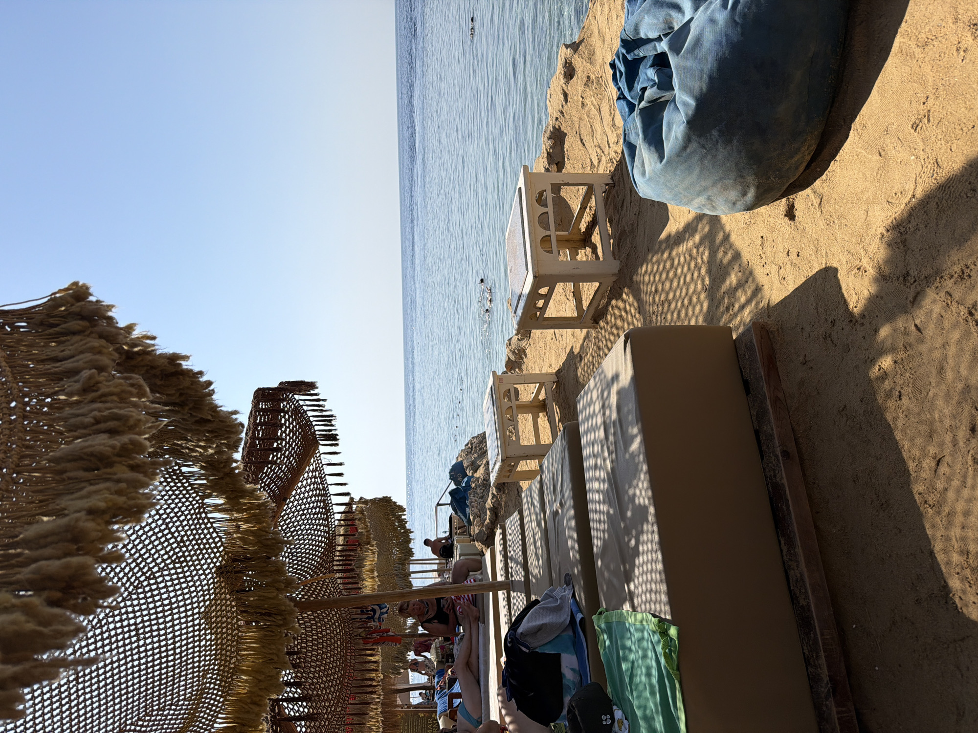 The image depicts a serene beach scene in Sharm El Sheikh, Egypt. In the foreground, several beige lounge chairs are arranged on the sandy shore, some adorned with green and blue towels. A large, dark blue beanbag chair rests to the right, casting a shadow on the sand. Behind the lounge chairs, small white tables with decorative cutouts sit on the sand, positioned near the water's edge. The background features the calm, turquoise waters of the Red Sea, with a clear blue sky above. A rocky shoreline separates the sand from the water. In the distance, a few people are visible in the water, some appearing to be swimming or snorkeling. The beach is shaded by straw umbrellas with tasseled fringes, creating a relaxed atmosphere. The overall impression is one of a tranquil vacation spot on a sunny day.