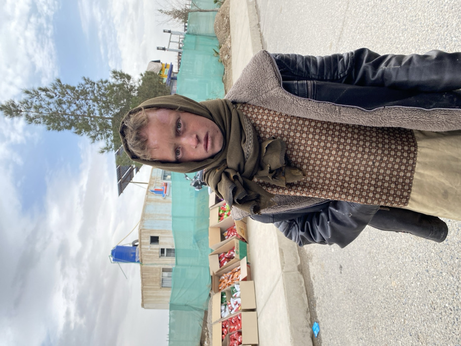 A young individual, appearing male, stands outdoors in Kandahar, Afghanistan, looking directly at the camera with a serious expression. They wear a dark olive-green headscarf, a brown patterned tunic-style garment, and a dark, possibly faux-leather, jacket with a shearling-lined collar. The headscarf and jacket show signs of wear and tear. In the mid-ground to the left, several open cardboard boxes filled with brightly colored packaged goods, likely snacks or candies, are arranged on a low surface, suggesting a roadside vendor setup. Behind this, a light-colored modular building or container structure is visible, topped with a blue water tank and an array of solar panels. A light-green mesh fence runs horizontally across the background, partially obscuring a motorcycle on the left and a dirt mound on the right. A tall, slender evergreen tree stands in the right background. The ground in the foreground is asphalt pavement. The sky is overcast with varying cloud cover.
