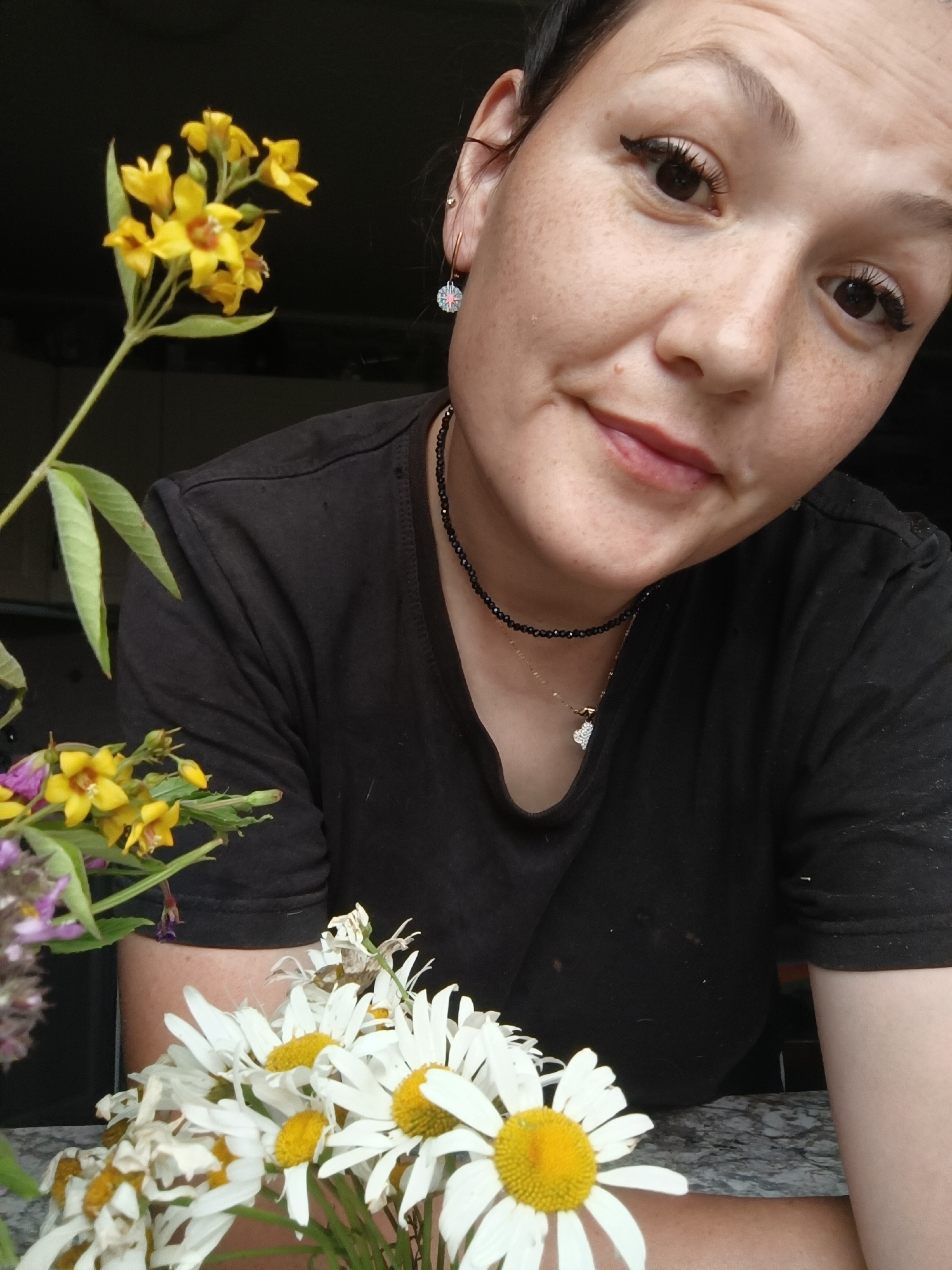 A close-up, high-angle, indoor selfie shows a young woman with freckles and dark hair pulled back. She is wearing a black t-shirt and a black beaded necklace with a small gold pendant. She is smiling slightly, with a hint of a blush on her cheeks. In the foreground, a bouquet of white daisies and yellow wildflowers sits on a speckled grey surface. The background is dark and out of focus, suggesting the photo was taken near a window or in low light. The location is not specified in Karsha, Russia, or any other city.