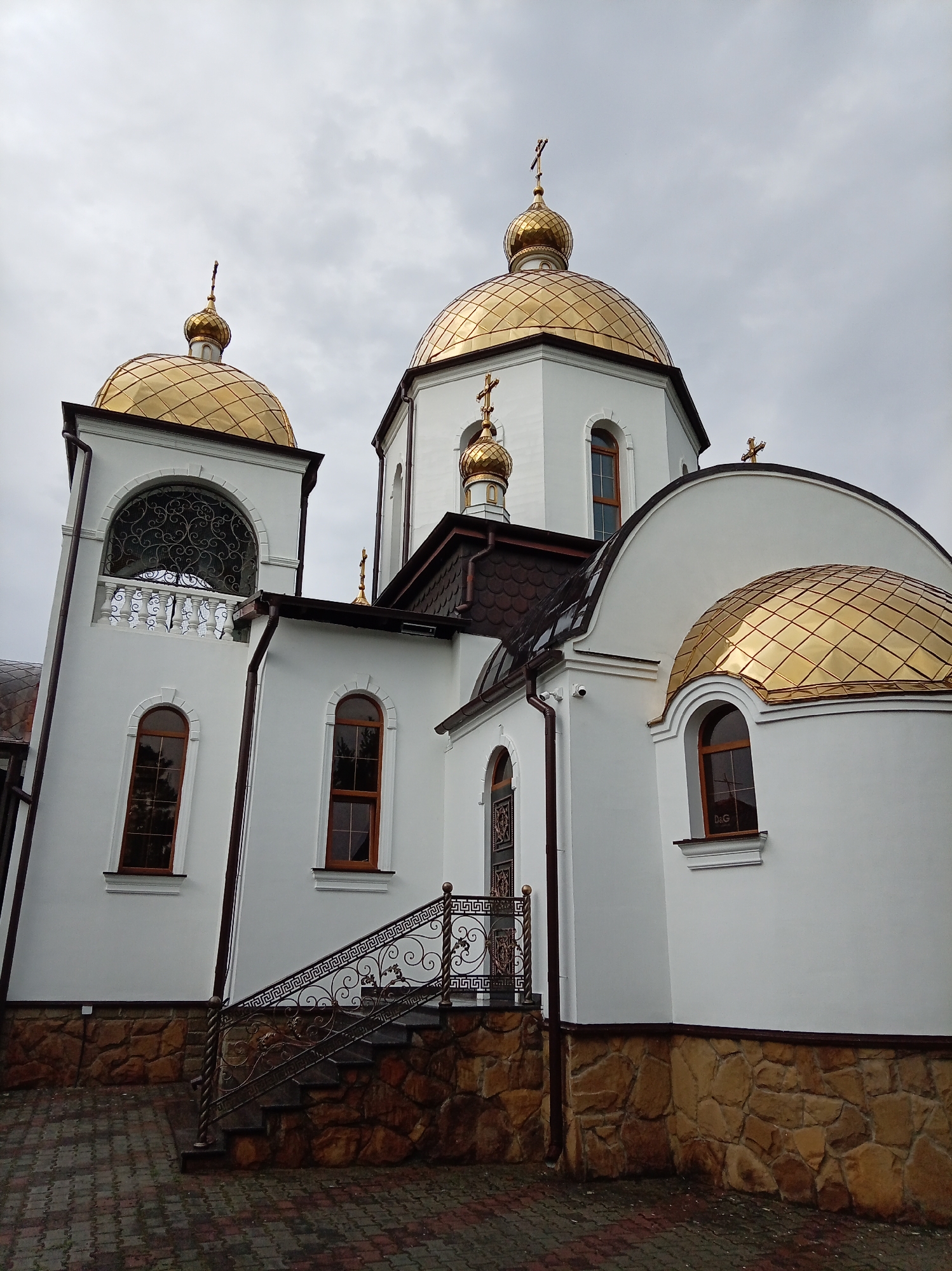 The image shows a close-up, low-angle view of the exterior of a white church with golden domes, located in Yessentuki, Russia. The church has several architectural features visible, including arched windows with dark frames, decorative metalwork on a balcony and a staircase railing, and a stone base along the lower portion of the building. The domes are a prominent feature, reflecting the light under a cloudy sky. The ground in front of the church is paved with brick pavers. There are no people or animals visible in the image. The overall scene is one of religious architecture under overcast weather conditions.