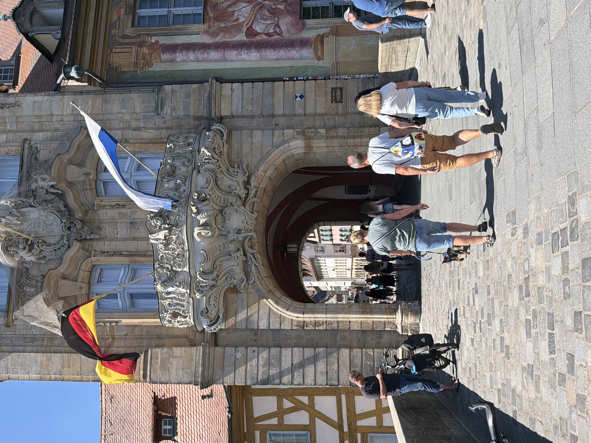 The image shows the historic Old Town Hall (Altes Rathaus) in Bamberg, Germany, which is situated on an island in the Regnitz River. The building is characterized by its ornate facade, featuring a large archway and balconies adorned with elaborate sculptures. Two flags are visible: a German flag and a blue and white flag, possibly representing Bavaria or the city of Bamberg.
In the foreground, several people are walking across a cobblestone bridge. A man in a dark t-shirt and blue jeans stands with a bicycle. Further ahead, a woman in a green t-shirt and denim shorts walks a small dog on a leash. Behind them, a couple walks arm-in-arm. The man wears a striped t-shirt and brown shorts, carrying a colorful backpack, while the woman wears a white t-shirt and blue pants. Other individuals are seen walking through the archway and in the distance. The environment is sunny with clear blue skies, and shadows are cast across the cobblestones. The architecture of the surrounding buildings, including half-timbered structures, is also visible.