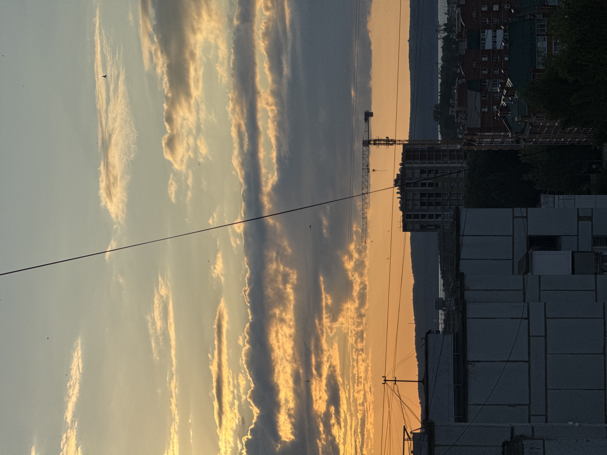 The image shows a cityscape at sunset, likely in Chaikovsky, Russia. The sky is filled with dramatic clouds tinged with orange and yellow from the setting sun. In the foreground, there are several buildings, including apartments and what appears to be a construction site with a crane. A prominent wire runs diagonally across the image, obscuring parts of the view. The overall impression is one of an urban environment at the end of the day, with the warm hues of the sunset contrasting with the dark silhouettes of the buildings and the sky.