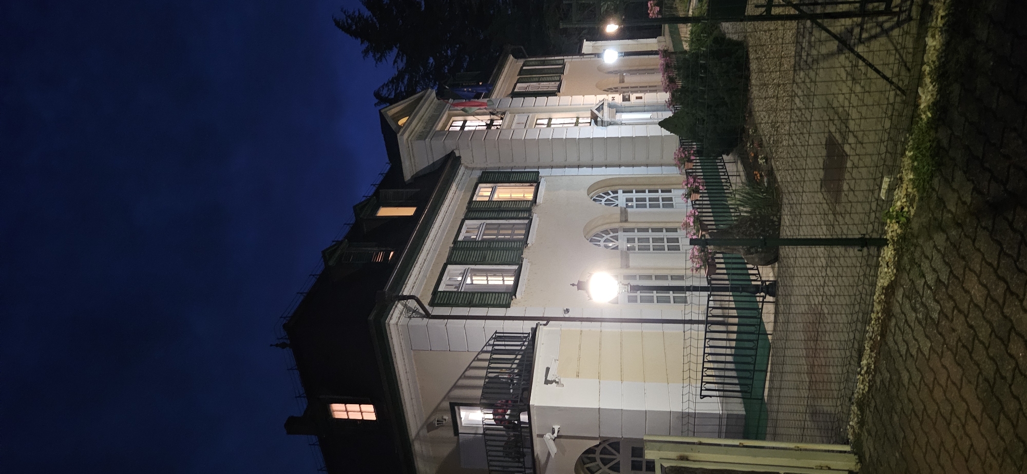The image depicts the exterior of a building in the city of Hévíz, Hungary, at dusk. The building appears to be a large, multi-story structure with a dark roof and cream-colored walls. Several windows are illuminated from within, casting a warm glow on the facade. Green shutters are visible on some of the windows. A balcony with a black railing extends from the upper level of the building.
In front of the building, there is a paved walkway or driveway, and a low black fence encloses a garden area. The garden contains several bushes, flower beds with pink flowers, and a prominent black lamppost with an illuminated globe. Another lamppost is visible further down the path. Two security cameras are mounted on the exterior wall of the building.
The sky above is a deep blue, indicative of twilight. Trees are visible in the background on the right side of the image. The overall scene suggests a well-maintained property, possibly a residential building, a diplomatic mission, or a commercial establishment, situated in a quiet, possibly suburban or park-like area of Hévíz. There are no people visible in the image.
