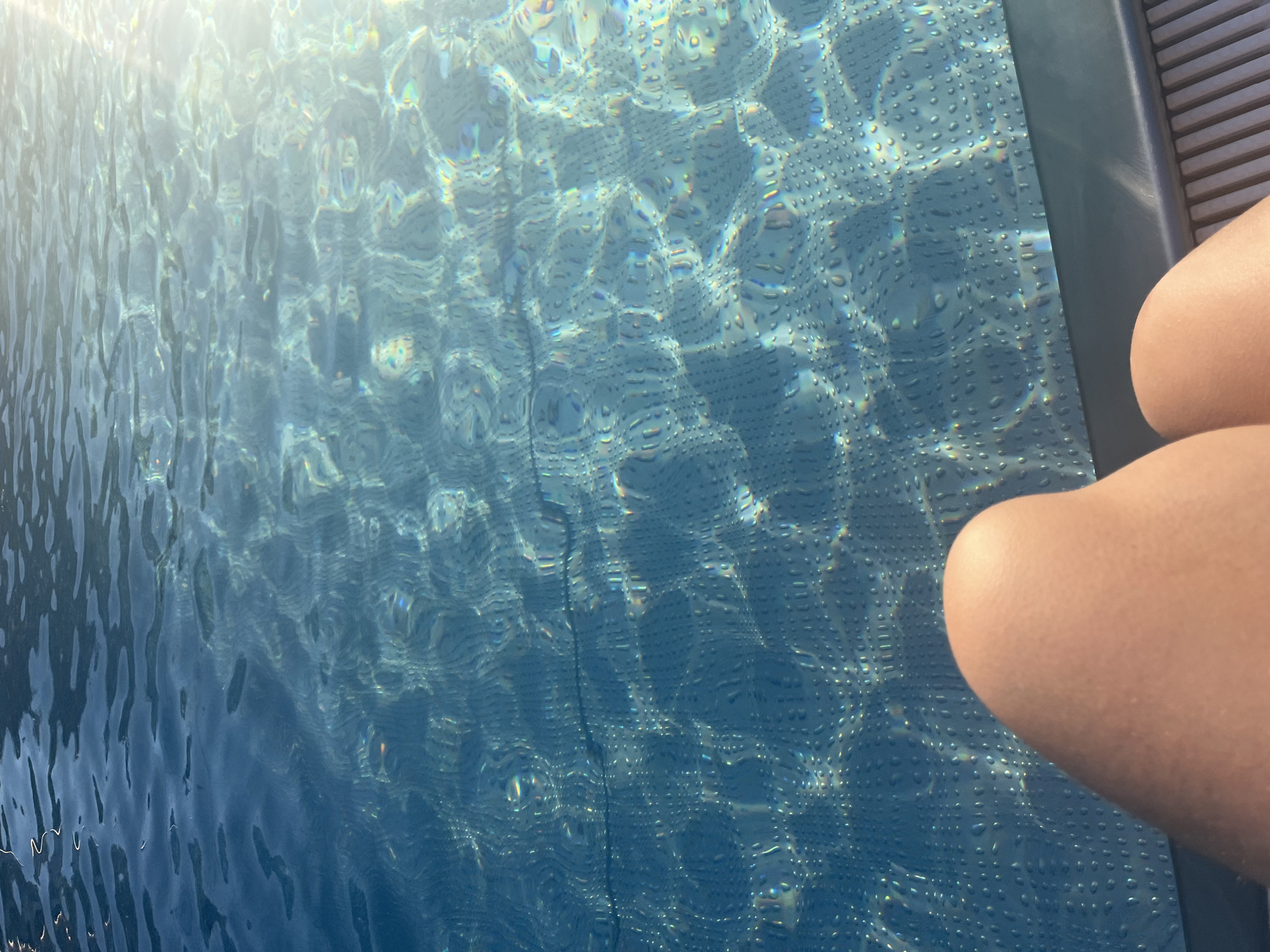 An overhead, close-up shot shows a person's thighs and knees at the bottom of the frame, positioned in front of a swimming pool. The pool water is a deep blue and is illuminated by sunlight, creating a shimmering effect with visible light reflections and refractions. The pool bottom is visible through the water, with a pattern of small, circular indentations. The edge of the pool, a dark blue coping, separates the water from a light-colored deck made of parallel wooden slats. The scene appears to be outdoors during the day, given the strong sunlight.