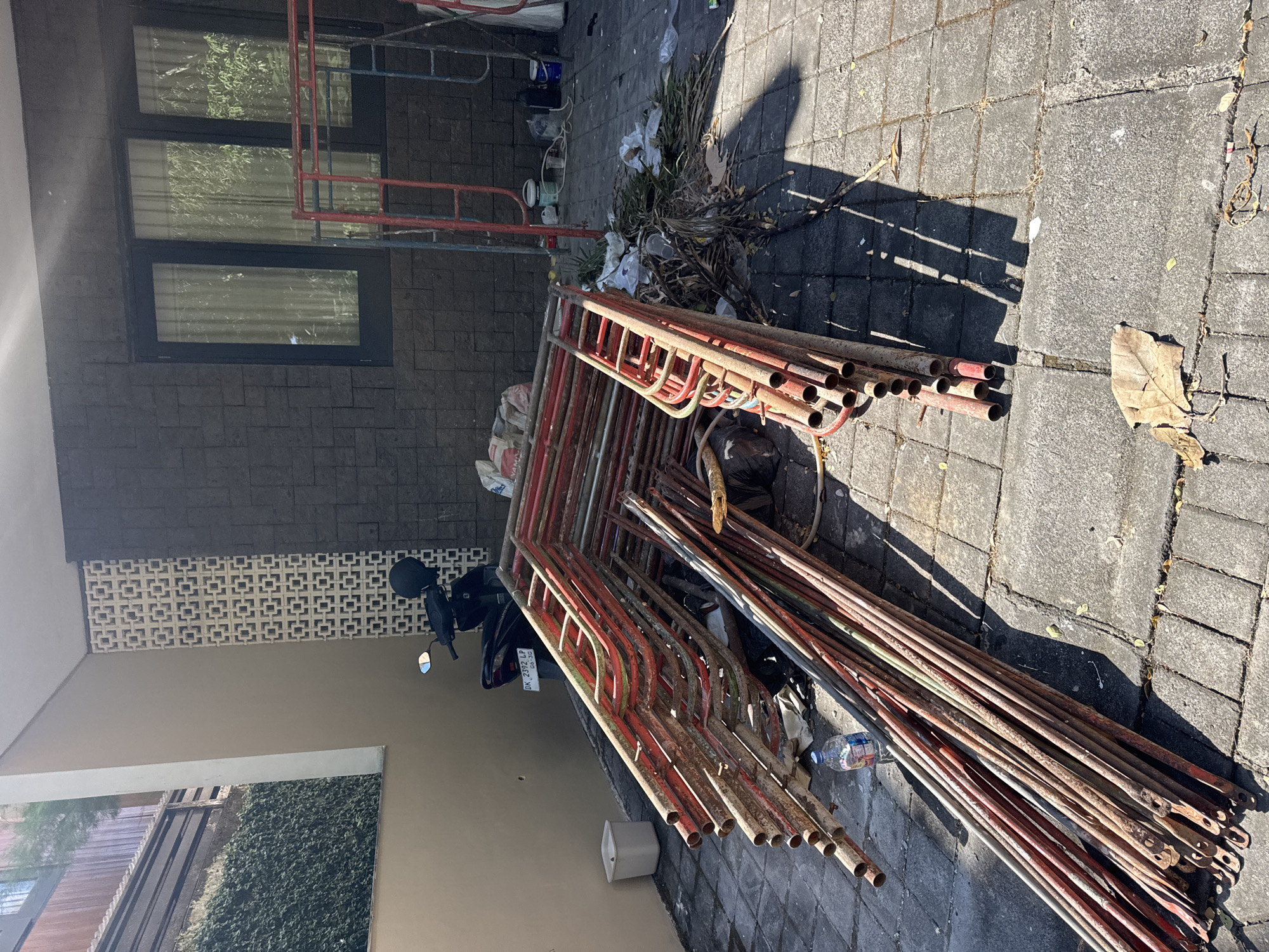 This image captures an outdoor, paved area adjacent to a building in Kuta Selatan, Indonesia, confirmed by the "DK" prefix on the visible motorcycle license plate (DK 2392 LP 08-30). The scene is illuminated by bright daylight, casting strong shadows from left to right.
The central foreground is dominated by a large pile of rusted metal scaffolding components. These consist of numerous red-painted frames and unpainted or weathered metal poles, stacked horizontally on grey interlocking concrete pavers. A plastic water bottle lies near the base of these poles. To the right of the stacked scaffolding, a scattered pile of debris includes green and dried plant matter, white plastic bags, and other litter.
In the mid-ground, a black scooter is parked facing left against a light beige wall section of the building. A black helmet rests on its seat. Above the scooter, the building features a decorative vertical panel composed of white patterned ventilation blocks. Further to the right, the building transitions to a dark grey textured stone or tile wall, in which three dark-framed windows with light-colored curtains are set. Several individual red and blue metal scaffolding frames stand upright or are partially assembled in front of this dark wall section. Near these, various items are visible on the pavement, including a white plastic container, several paint cans (blue and green), and a black container.
The paved ground shows signs of wear and dirt. A large, dry brown leaf is prominently featured in the lower foreground. To the far left, a lush green hedge and a dark wooden fence or barrier are visible, suggesting an adjacent property or landscaped area. A vertical white column delineates a section of the building. No people are present, and no active work or interaction is depicted.