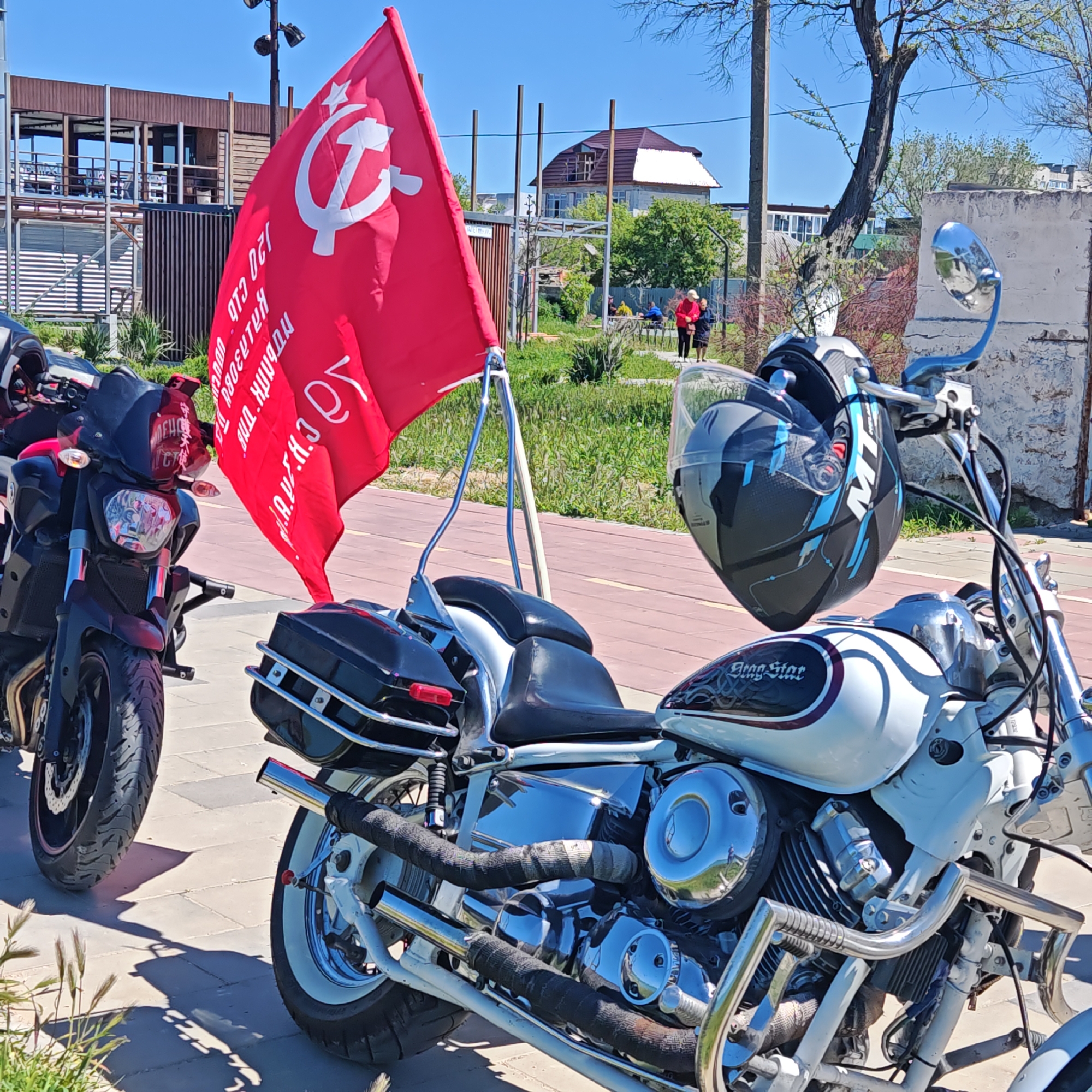 The image shows two motorcycles parked on a paved area, possibly a sidewalk or plaza, in the city of городское поселение Щелкино, Ukraine. The motorcycle in the foreground is white with chrome accents and a black helmet hanging from the handlebars. The helmet is mostly black with blue designs. The motorcycle has the words "Drag Star" on the fuel tank. To the left is a dark-colored motorcycle with a windscreen. A red flag featuring a white hammer and sickle emblem, associated with communism, is attached to the rear of the white motorcycle. The text on the flag is in a foreign language. In the background, there's a structure that appears to be a covered seating area and a building. People are visible in the background. The sky is clear and blue. A tree is visible to the right, and vegetation and grass are present.