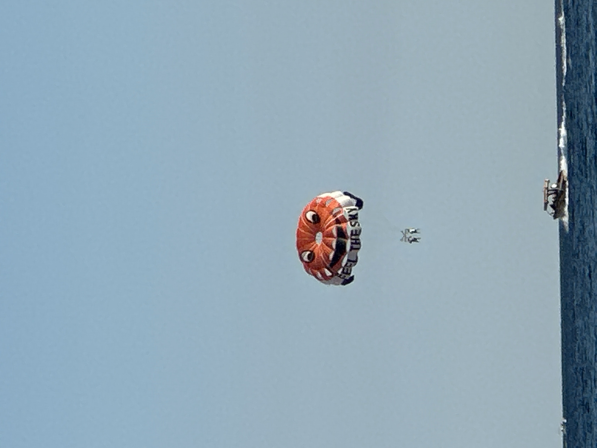 The image captures a clear sky over the sea in Boreti, Montenegro. A brightly colored parasail, designed to resemble a cartoon face with large eyes and a smiling mouth, is visible in the upper part of the frame. The parasail has text that reads "FEEL THE SKY". Two people are suspended beneath the parasail, enjoying the activity. In the lower portion of the image, a small motorboat is traversing the blue water, leaving a white wake. The sea is calm with some waves visible.
