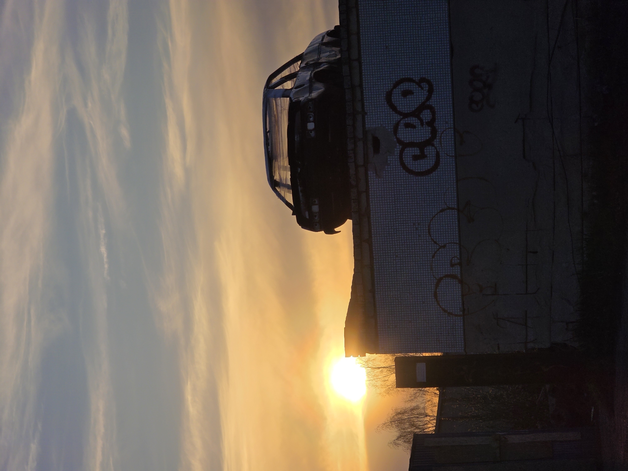 A rotated, low-angle shot reveals a landscape featuring a car perched atop a building near the city of Unknown, Unknown. The backdrop showcases a sky painted with the hues of a sunset, casting a warm glow around the sun's position. The building's wall is adorned with graffiti, juxtaposed against the modern vehicle. Sparse tree branches peek behind the structure, adding a hint of nature to the urban scene.