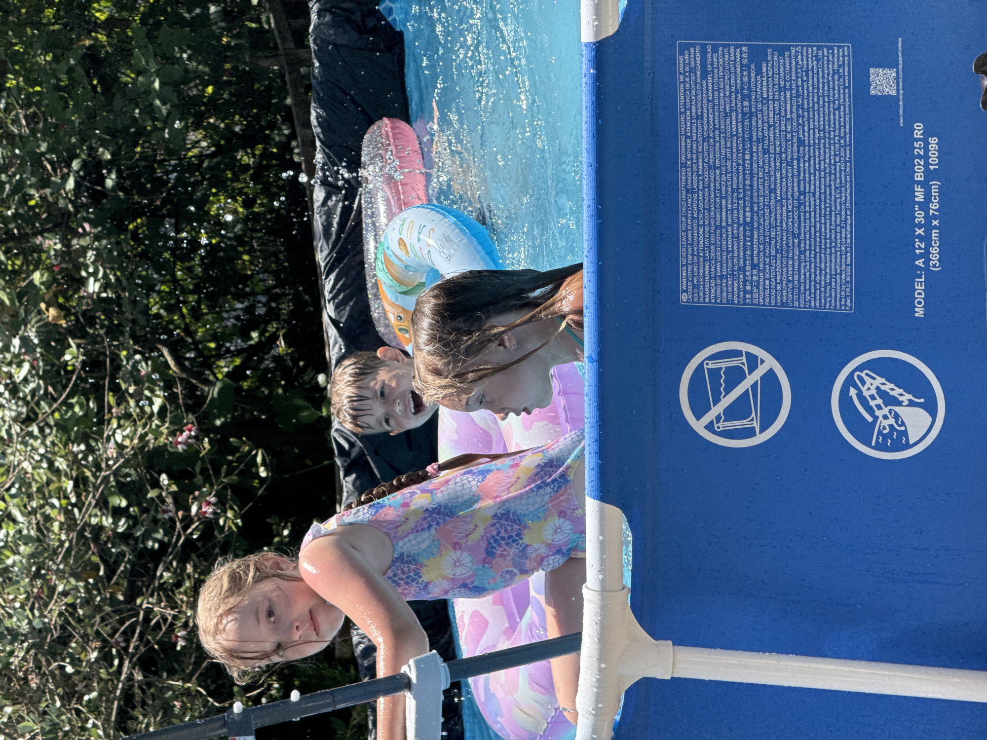 The image shows three children playing in a pool. A young girl in a colorful swimsuit is in the foreground, holding onto a pole. Behind her, a boy with light brown hair is smiling and looking at the camera. To the right, another girl with wet brown hair is looking to the side. Two inflatable rings, one pink and one with a colorful pattern, are floating in the water. The background is a blur of green foliage. The bottom of the image shows part of the pool structure with warning labels. The overall scene depicts a sunny day of summer recreation. The location is identified as Sochi, Russia.