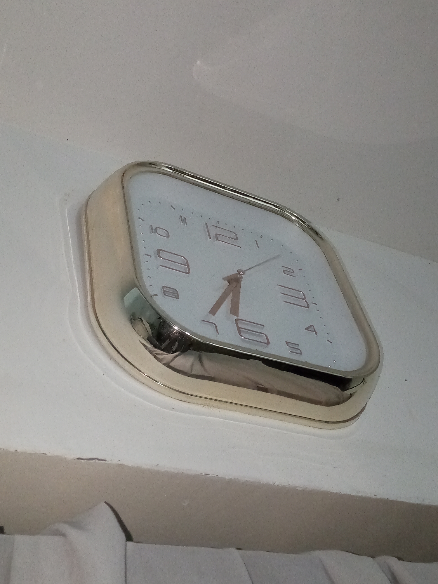 A low-angle close-up shows a gold-rimmed, white-faced wall clock with a slightly rounded, rectangular shape. The clock face displays the numbers 1 through 12 in a modern, sans-serif font. The hour hand points just past 6, and the minute hand is nearing 2, suggesting a time around 6:10. The clock is mounted on a white wall or surface. A partially visible white object, possibly fabric, is at the bottom of the frame. The scene appears to be indoors and possibly located in Davao City, Philippines, based on the request, but no specific geographic markers are visible in the image.