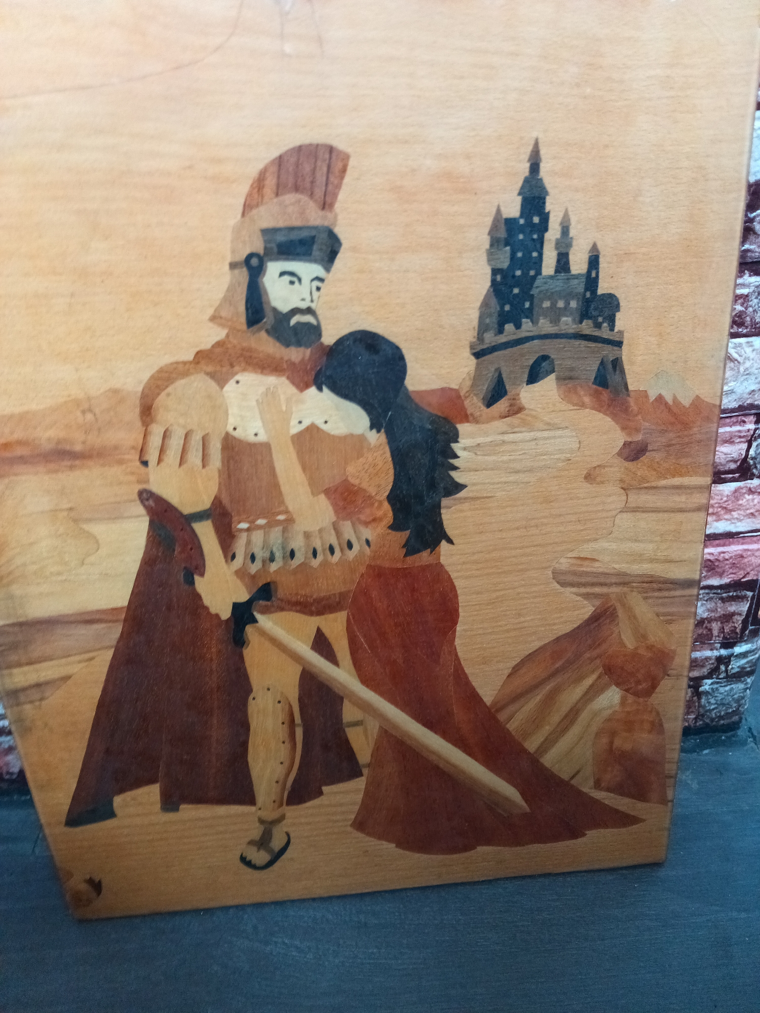 This vertically oriented wood inlay artwork, located in the city of Unknown, Unknown, depicts a stylized scene with two figures and a distant castle. On the left, a male figure stands, facing right, rendered in various shades of brown wood, with a prominent dark beard and mustache. He wears a helmet with a crest, light-colored puffy sleeves, a darker tunic with light trim, dark greaves with circular embellishments, and simple sandals. A dark reddish-brown cape drapes over his left side. His right hand holds a light-bladed sword with a dark hilt, pointing diagonally downward and right. His left arm is bent towards the female figure. On the right, a female figure stands, facing left, leaning into the male figure, her head resting near his chest. She has long, dark black hair and wears a reddish-brown long gown with a lighter bodice. Her hands are raised towards her face or the man's chest. In the upper right background, a multi-towered, dark blue-gray castle with visible windows is perched atop a brown, arched hill. The landscape is composed of varying light and dark brown wooden plains and undulating hills, with a lighter, path-like element in the mid-ground. The entire composition utilizes natural wood tones, giving it a rustic, artistic quality.