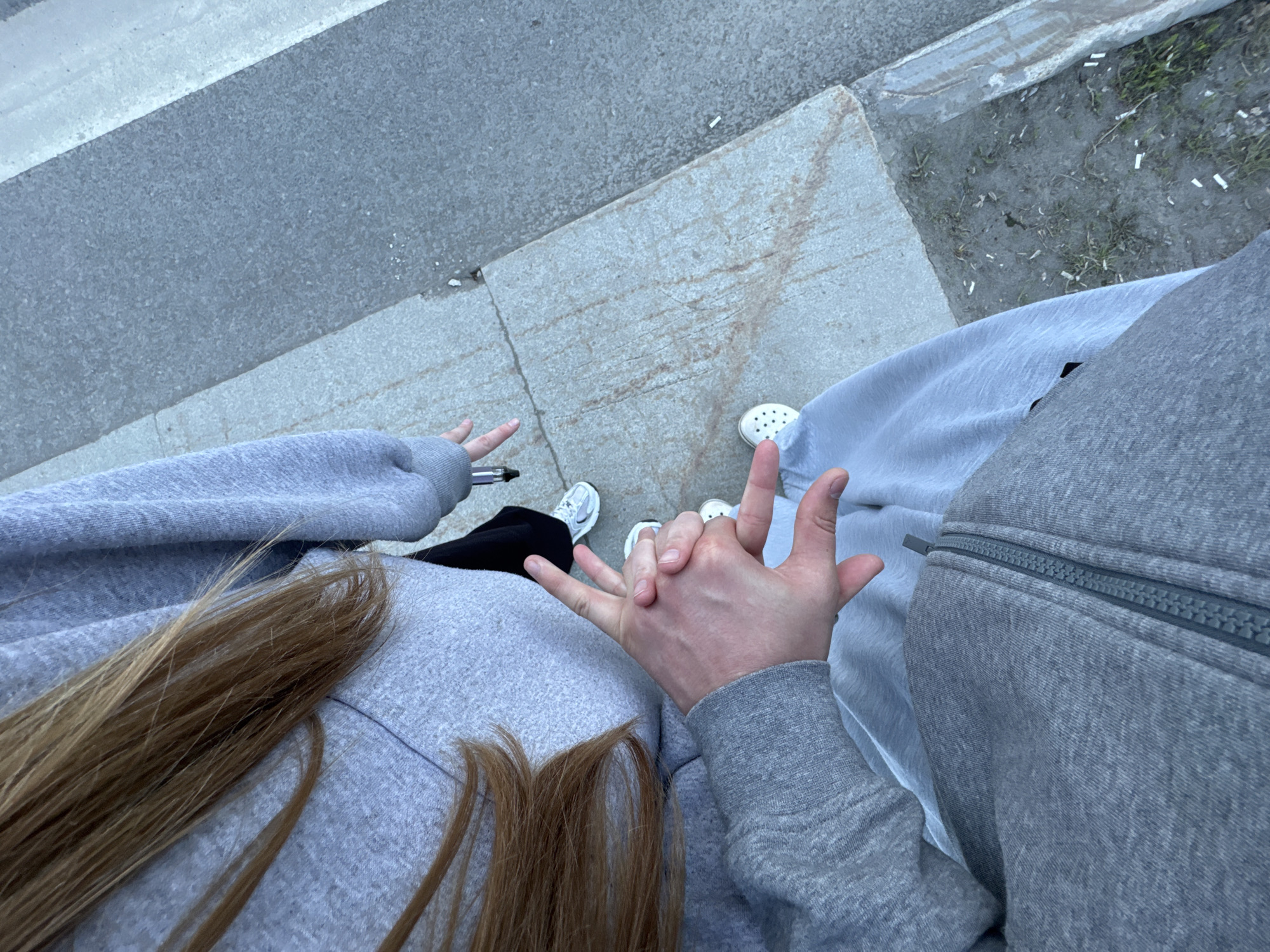 The image depicts a view from above, capturing two individuals standing on a sidewalk constructed from gray concrete slabs. The location may be in Perm, Russia. The slabs border an area with loose dirt, sparse vegetation, and discarded cigarette butts.
The two people are primarily visible from the torso down. The person on the left wears a gray sweatshirt, black pants, and white sneakers with gray accents. They hold an electronic cigarette or vape pen in their hand. Long, brown hair falls across their shoulder. The person on the right is clad in a gray zip-up sweatshirt and light-colored pants, with their footwear appearing to be white Crocs. Their hands are interlocked, with fingers extended in what appears to be a casual hand gesture or a shared signal.