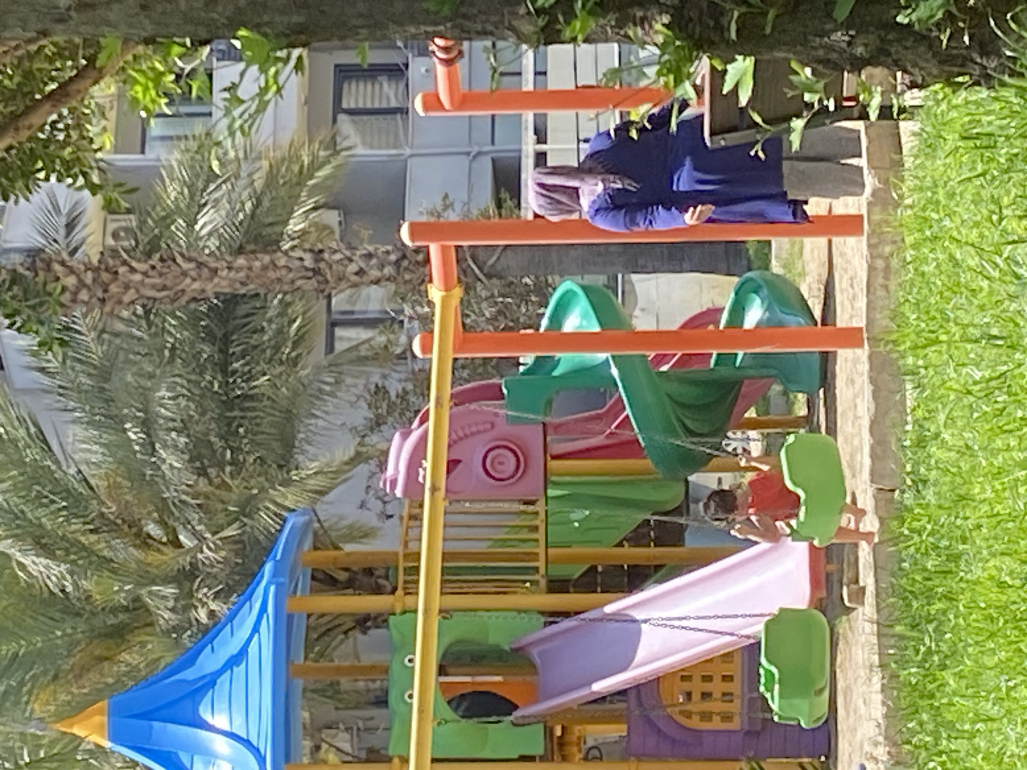 A colorful playground structure stands in Alanya, Turkey, surrounded by a palm tree on the left and green grass on the right. A woman in a blue dress and head covering stands near the top of the playground. A child in a red shirt sits on a swing. A multi-story building with windows is visible in the background.