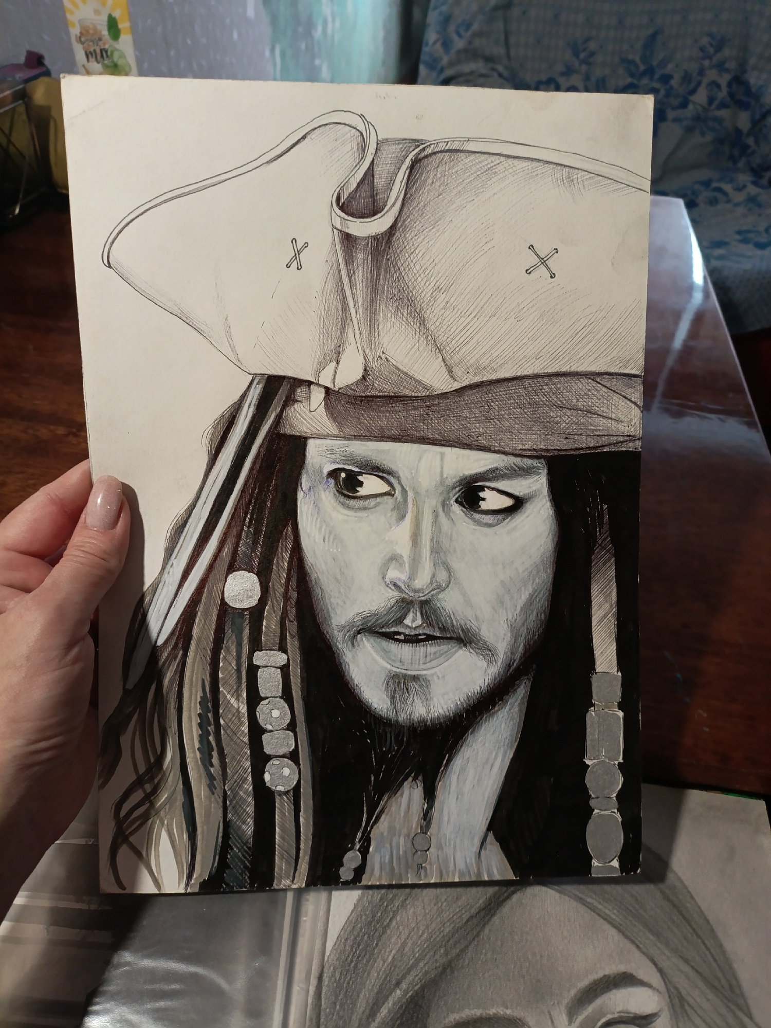 A close-up, eye-level, portrait-style photograph shows a drawing of Captain Jack Sparrow. The drawing is rendered in black and white with subtle gray and white shading. The subject wears a tri-cornered hat and has dark, braided hair adorned with beads. His eyes are wide and looking to the right, and he has a goatee. The drawing is on a white piece of paper, held at the left by a person's hand, revealing a painted fingernail. The background is blurred, showing hints of furniture and possibly a patterned fabric. The drawing appears to be on a table, with another drawing visible underneath it. The lighting is consistent, highlighting the details of the drawing.