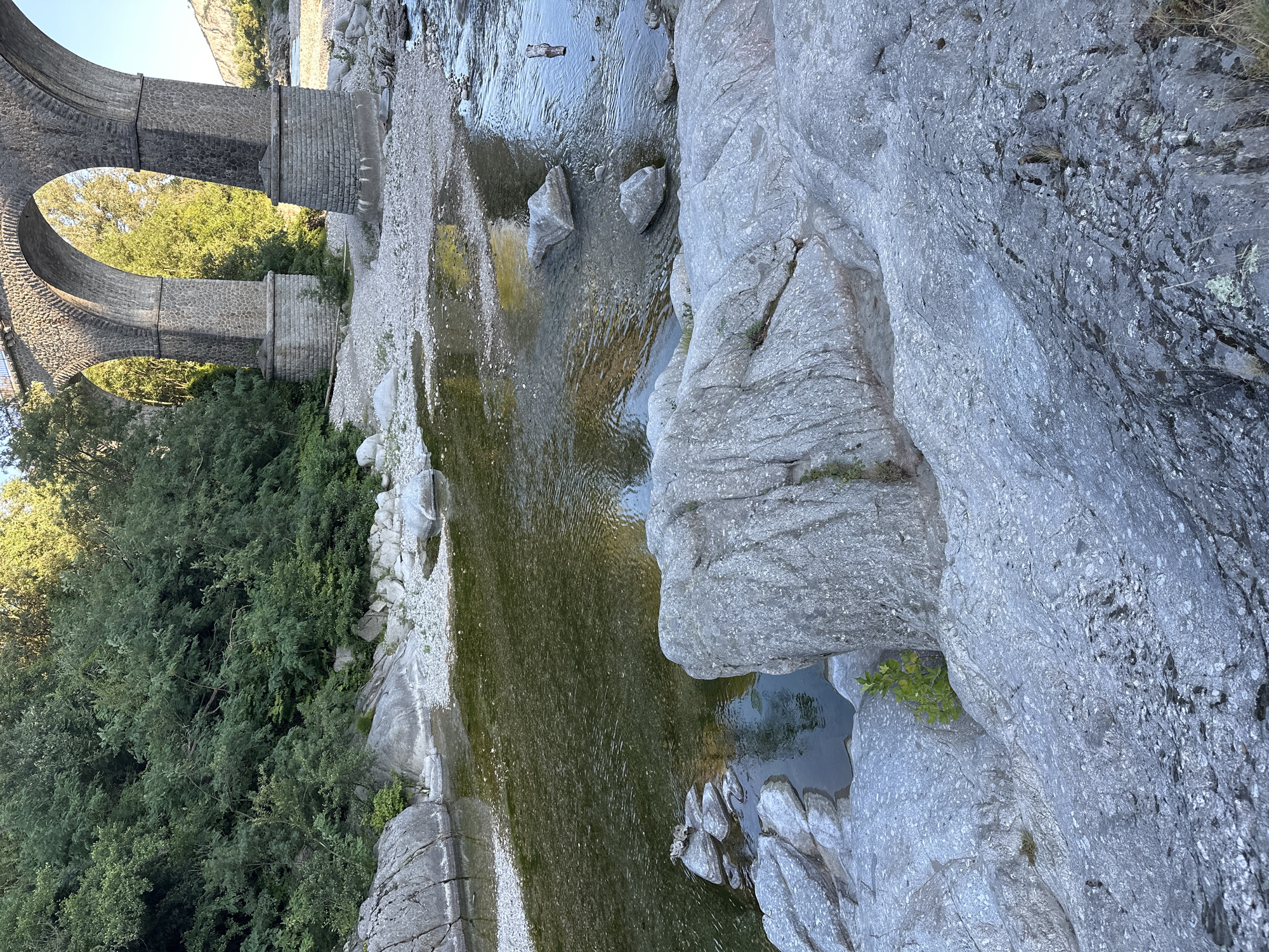 The image shows the rocky banks of the Gardon River in Anduze, France, beneath a stone arch viaduct. The water is shallow and clear, with sunlight reflecting off its surface. Large, smooth grey rocks are prominent in the foreground, with smaller pebbles and a gravelly shore extending towards the water's edge. Lush green trees and vegetation line the opposite bank, climbing the hillside. The viaduct, constructed from large stone blocks, has multiple arches and towers supported by substantial pillars. The overall scene suggests a natural, somewhat arid landscape with a historical man-made structure.
