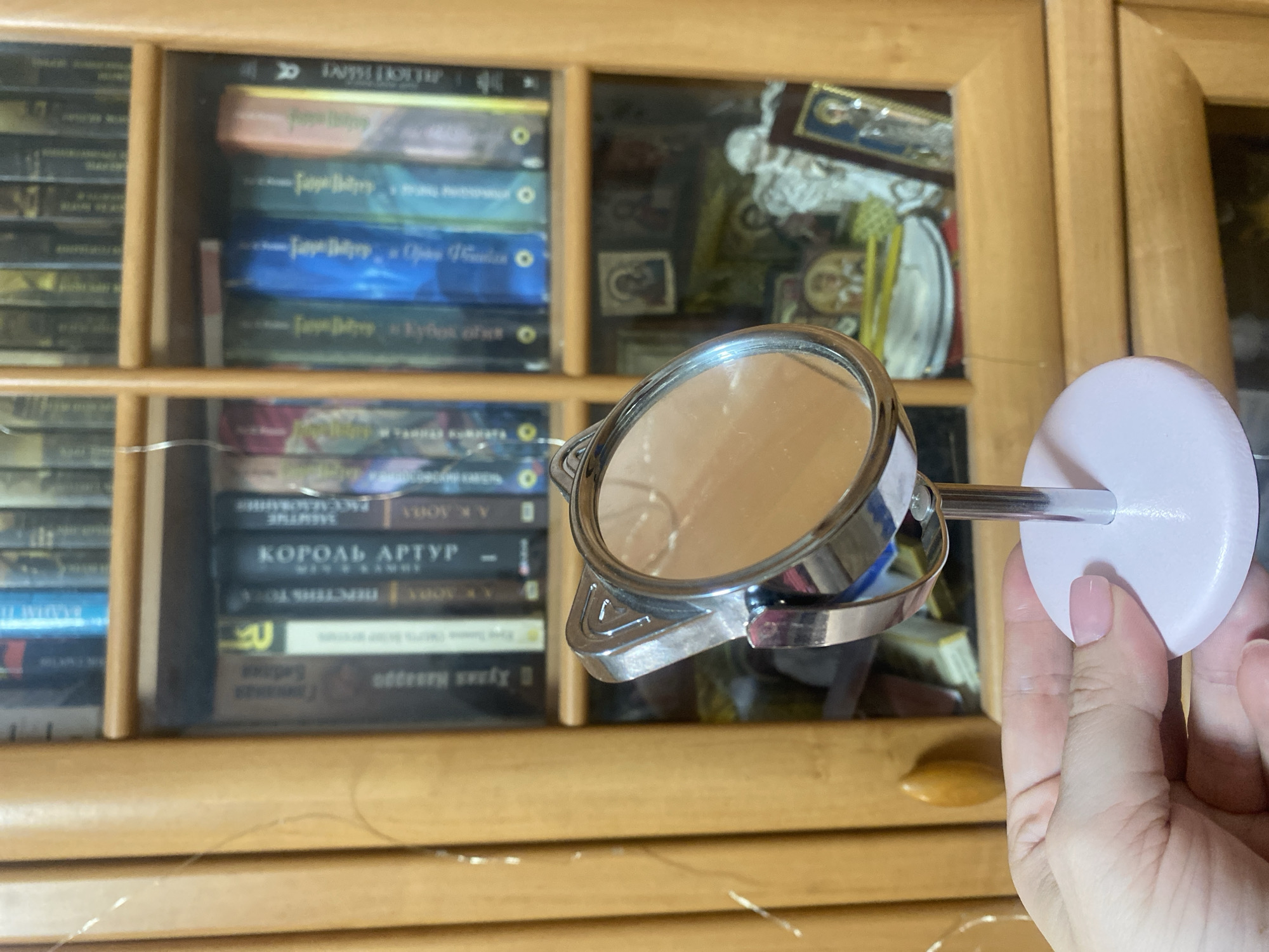A person's hand, with neatly manicured light pink fingernails, holds a small, round makeup mirror with a silver frame shaped like cat ears. The mirror stands on a slender silver rod attached to a light pink circular base. In the background, a wooden bookshelf is filled with books and religious icons. The bookshelf appears to be located in a home in Chornomorsk, Ukraine.