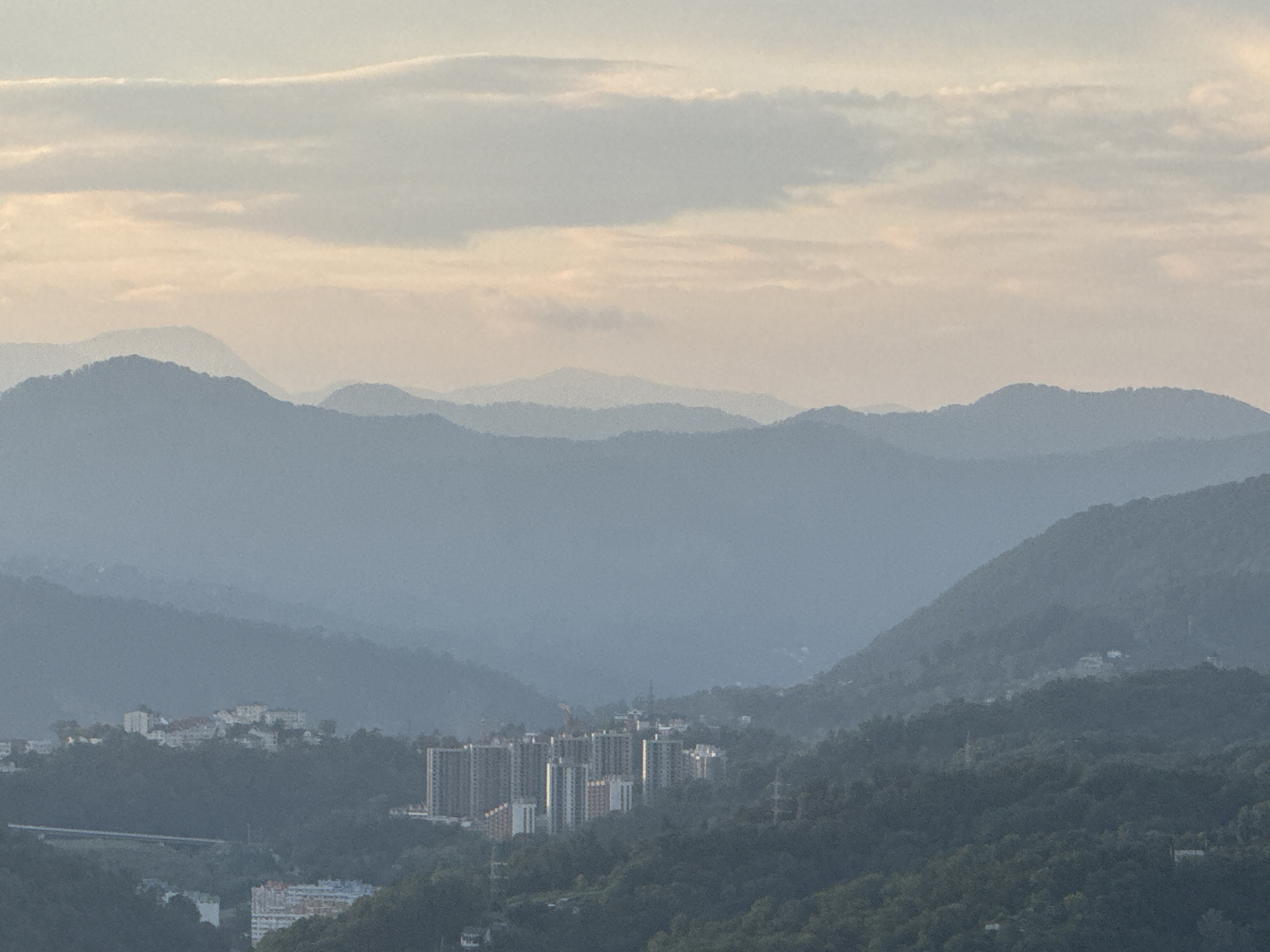 This is a wide-angle, aerial photograph of a valley with layered mountains and a city nestled within. The sky is overcast with soft, pale clouds, tinged with a peachy-pink hue near the horizon, suggesting either dawn or dusk.
In the foreground, lush green trees cover the slopes of the mountains. The middle ground reveals a collection of buildings, including a prominent cluster of modern, multi-story residential buildings with balconies and a few smaller, white structures. A linear road or bridge is visible in the lower left quadrant. Further into the valley, more residential buildings and infrastructure are scattered across the landscape, partially obscured by the misty atmosphere.
The background is dominated by numerous mountain ranges that recede into the distance, their outlines softened by the haze. The closest mountains are a deep blue-grey, while the further ones appear progressively lighter and more muted in color, creating a sense of depth.
The scene appears to be a view from a higher vantage point overlooking a part of the city of Sochi, Russia. The architectural style of the prominent buildings suggests a developed urban area within a mountainous region. The overall atmosphere is serene and tranquil, with a focus on the natural landscape interacting with human settlement. There are no visible people in the image.