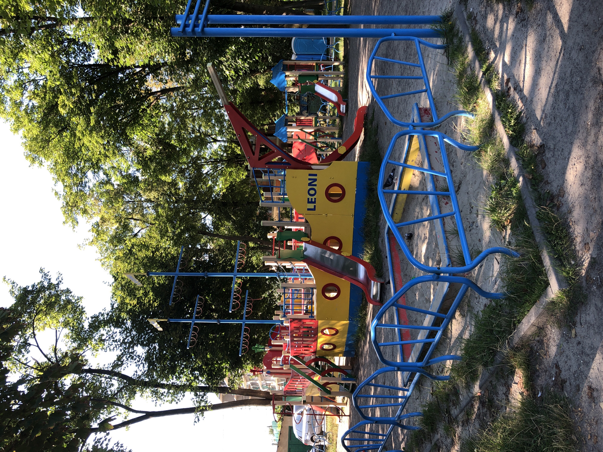 The image depicts a vibrant and colorful children's playground designed to resemble a pirate ship. The main structure is a large, yellow and blue ship with a red slide and various play features. The playground is situated outdoors, surrounded by lush green trees that provide shade. Sunlight filters through the leaves, casting dappled shadows on the sandy ground. The scene appears to be set during the daytime, likely on a clear, sunny day.
Prominent objects include the ship-themed playground equipment, made of durable, brightly colored materials like plastic and metal. There are blue metal railings and supports, a red and yellow ship body, and a prominent red slide. The ship has masts with what appear to be decorative flags. Scattered throughout the playground are smaller play structures, including swings and climbing elements. The ground is a mix of sand and grass, with a concrete border running along the edge.
While no people are visible in the image, the playground is clearly designed for children's recreation. The overall mood is one of fun and adventure. The text "LEONI" is visible in black lettering on the side of the yellow portion of the ship structure. The location is identified as Kolomyia, Ukraine.