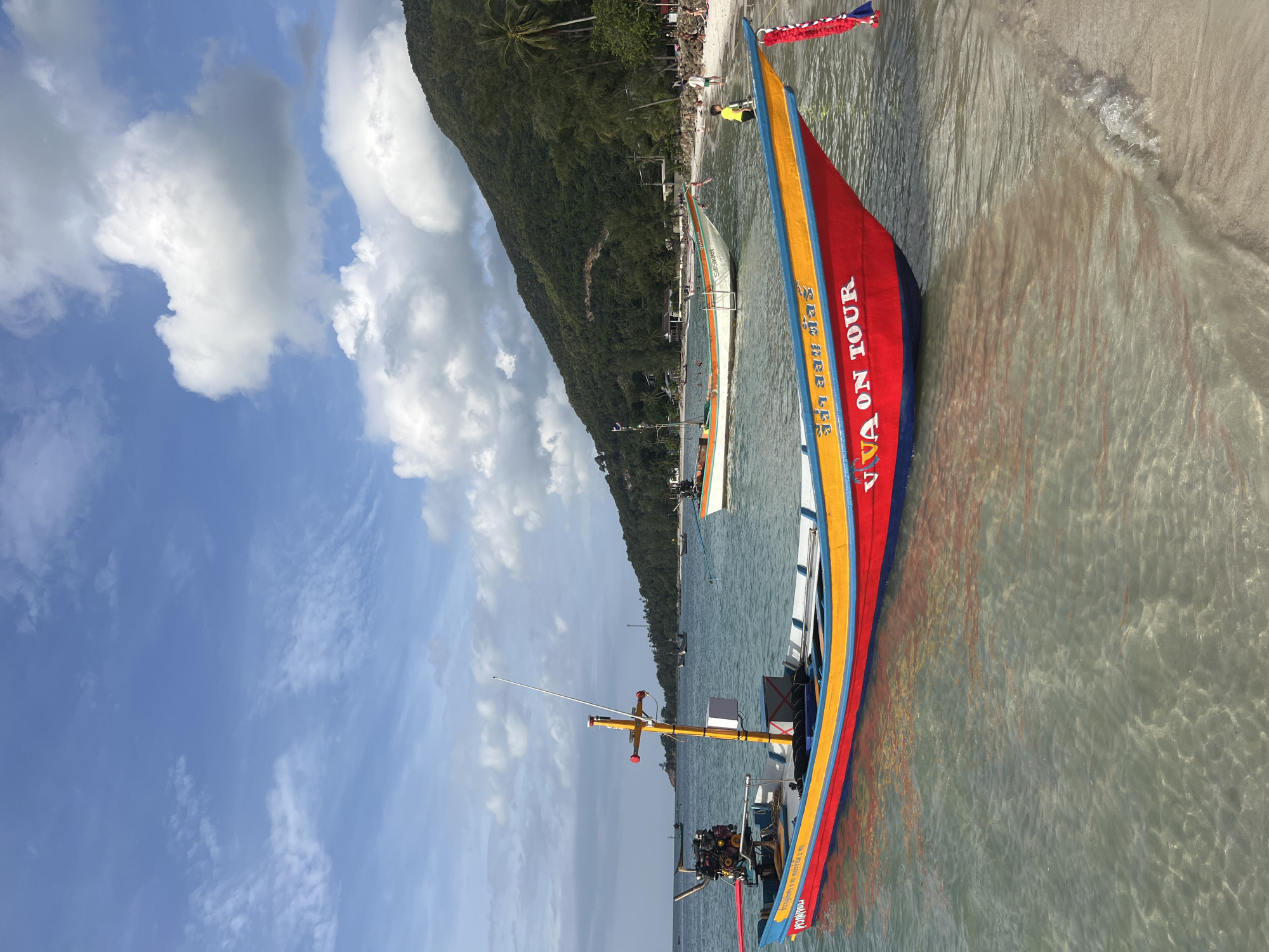 A brightly colored longtail boat, painted red, yellow, and blue, is anchored in shallow, clear water close to a sandy beach. The words "VIVA ON TOUR" are visible in white lettering on the side of the boat. Other longtail boats are moored in the background, and beyond them, a lush, green, tree-covered hillside slopes down to the shore. Several people are visible on the beach, some walking and others relaxing. The sky is blue with scattered white clouds. The scene depicts a tropical beach setting.