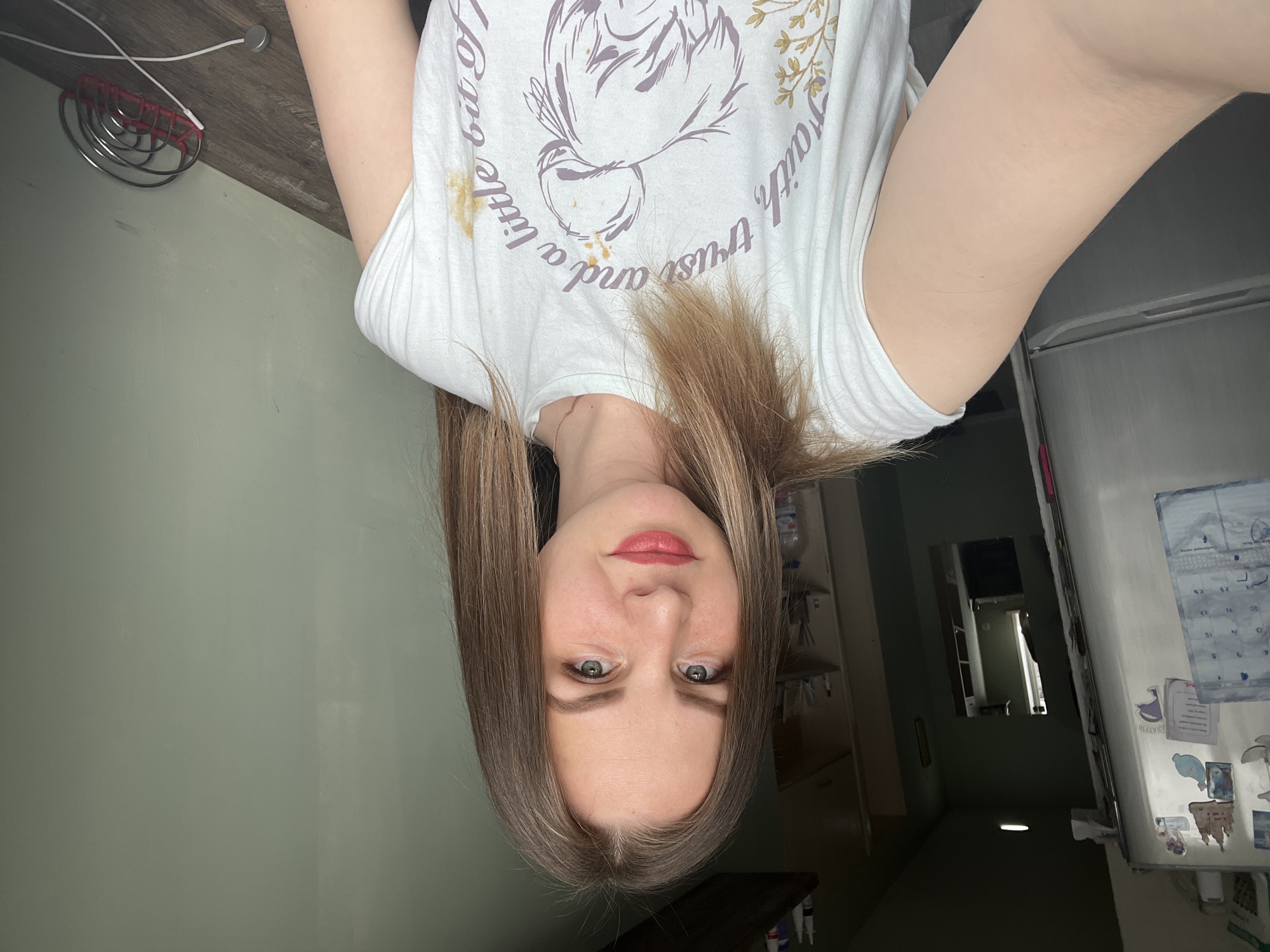 A medium-shot selfie of a Caucasian woman with medium-length brown hair, wearing a light-colored t-shirt with a graphic design. She is facing the camera with a neutral expression. The background includes a green wall, a refrigerator with magnets and a calendar, and a hallway leading to another room. The location is not identifiable as Izhevsk, Russia.