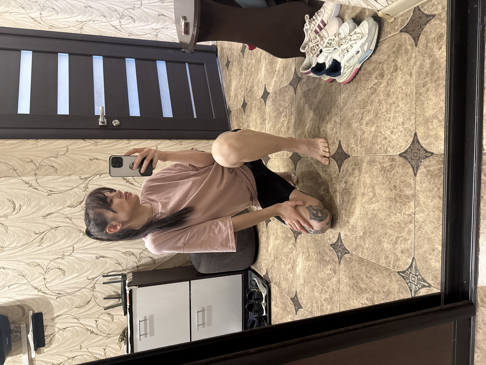 A mirror image shows a woman taking a selfie indoors. The woman has dark hair with bangs and is wearing a pink t-shirt and black shorts. She is kneeling on a gray cushion. Her left leg is extended, and her right leg is bent with her hand on her knee, which has a tattoo. She is holding a phone in her right hand, taking a picture of herself in the mirror. The mirror reflects a closed dark door with horizontal light panels. A small cabinet is to the left of the woman, and a pair of shoes are on top of a round brown table to the right. The floor is tiled with beige patterned tiles. The city of Rostov-on-Don, Russia, is not discernible from the image.