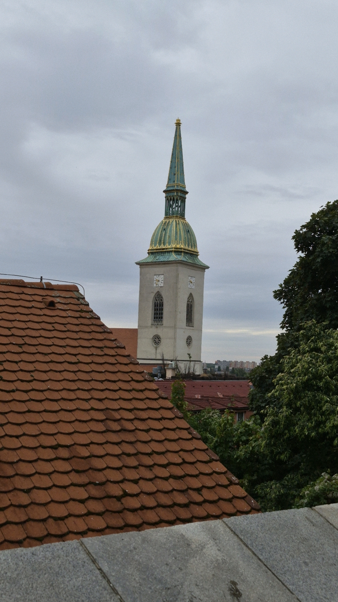 This is a vertical, eye-level shot of the St. Martin's Cathedral spire in Bratislava, Slovakia, framed by a foreground of terracotta roof tiles and a grey stone wall. The cathedral, a tall, white structure with arched windows and a green, gold-adorned spire, is situated in the middle ground, slightly to the right of center. The spire's tip pierces a cloudy, grey sky. To the right of the cathedral, lush green trees obscure a portion of the background, which features a distant cityscape of buildings. The sky is overcast, suggesting a cloudy or possibly rainy day. There are no people visible in the image.