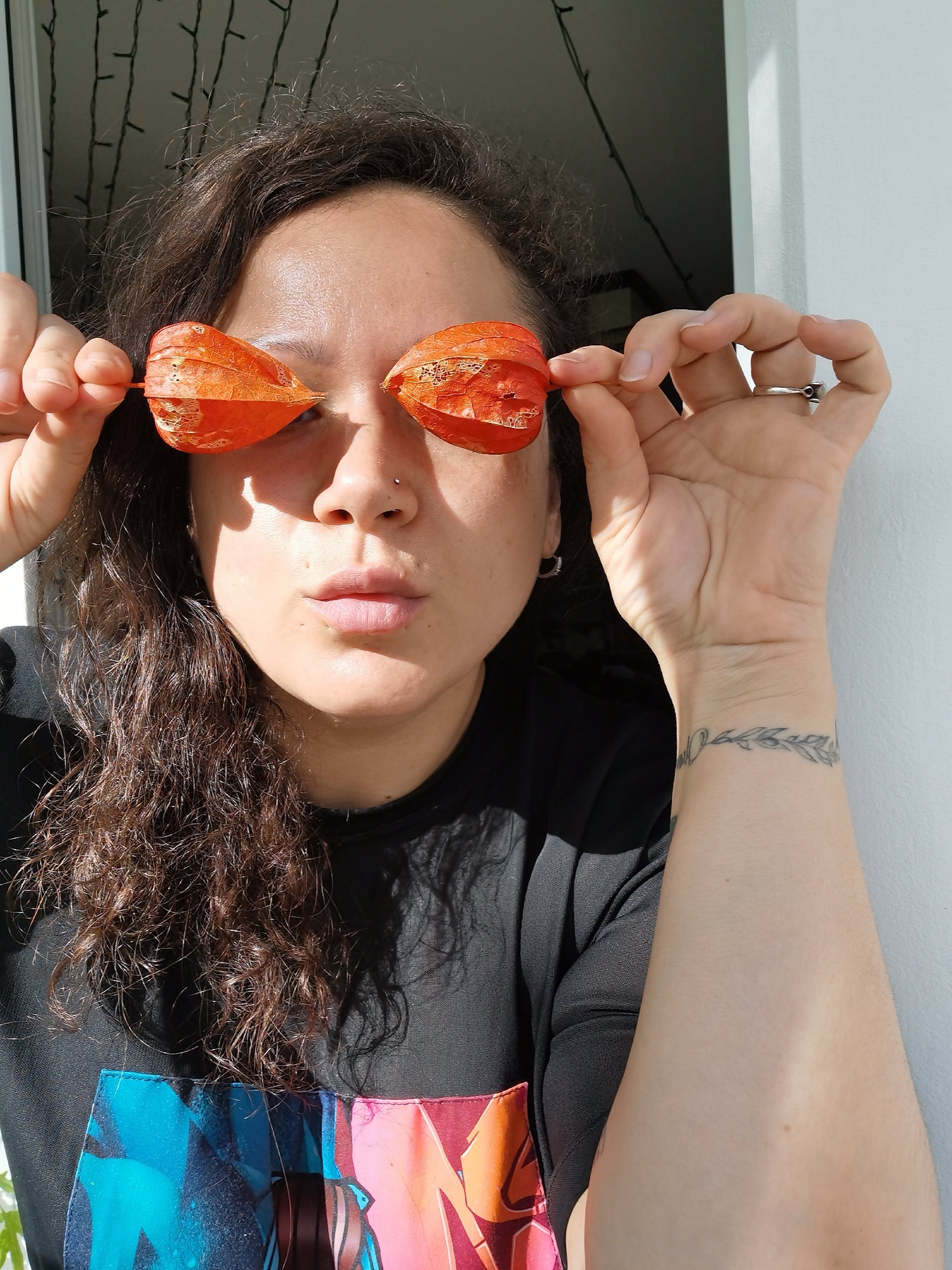 A woman is indoors, likely near a window or doorway based on the natural light and the glimpse of the outside. She is holding two bright orange, lantern-shaped objects (possibly Physalis fruit) over her eyes, mimicking sunglasses. She has dark, curly hair and a fair complexion. She has a nose piercing and a silver ring on her right hand. There is a tattoo on her left wrist. She is wearing a black t-shirt with a colorful graphic print on the bottom. The background shows a white door frame or wall and a dimly lit area with faint hints of vertical lines, possibly representing window blinds or a fence.
I cannot confirm the exact location in Chaikovsky, Russia based on the image alone.
