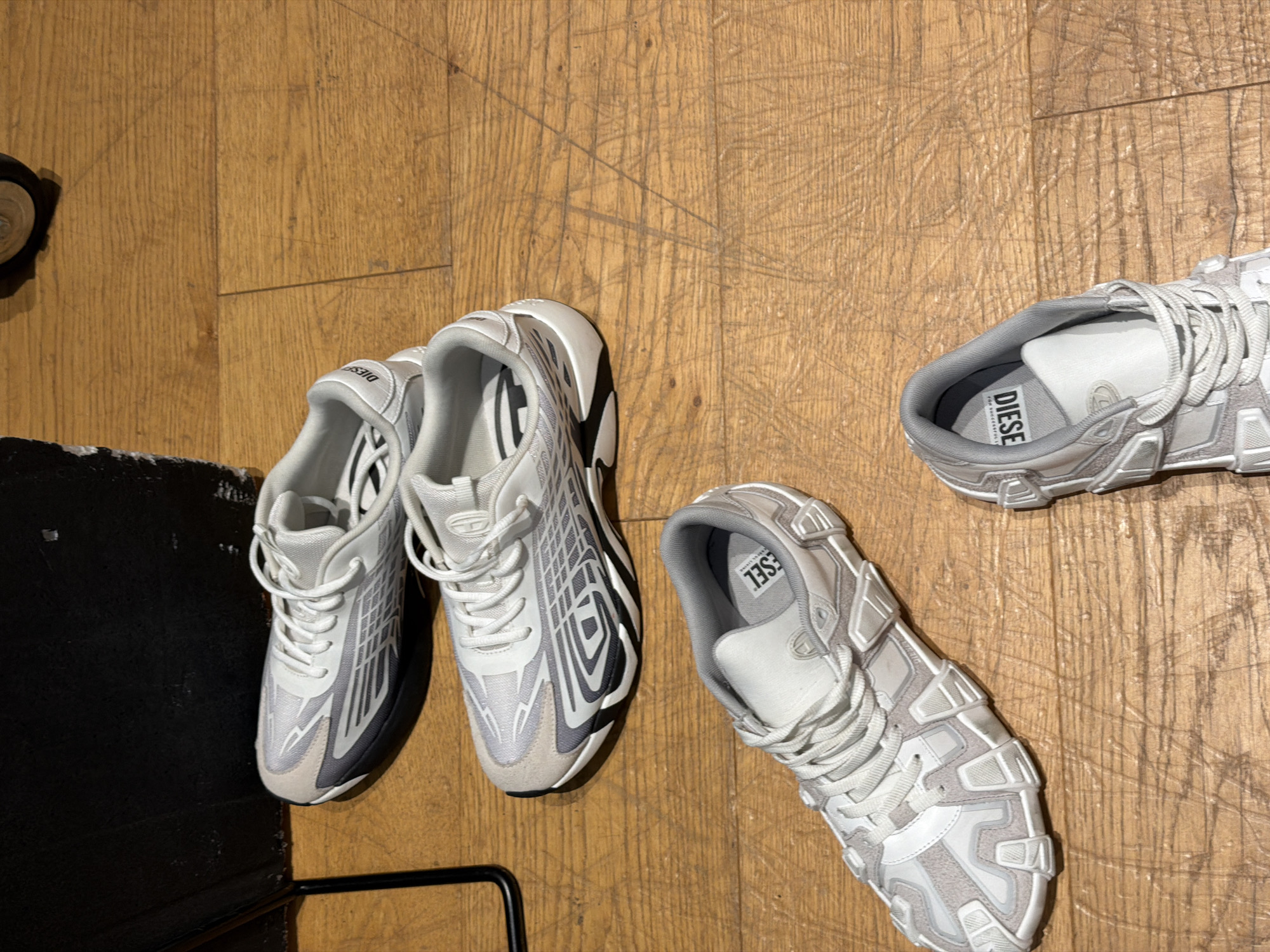 The image depicts two pairs of white Diesel brand sneakers on a wooden floor. The first pair, located to the left, is placed near a black object, possibly a piece of furniture. The second pair is positioned further to the right. The environment appears to be indoors. No people or actions are visible. The city of Mirabella, Italy, is not evident from the visual information.