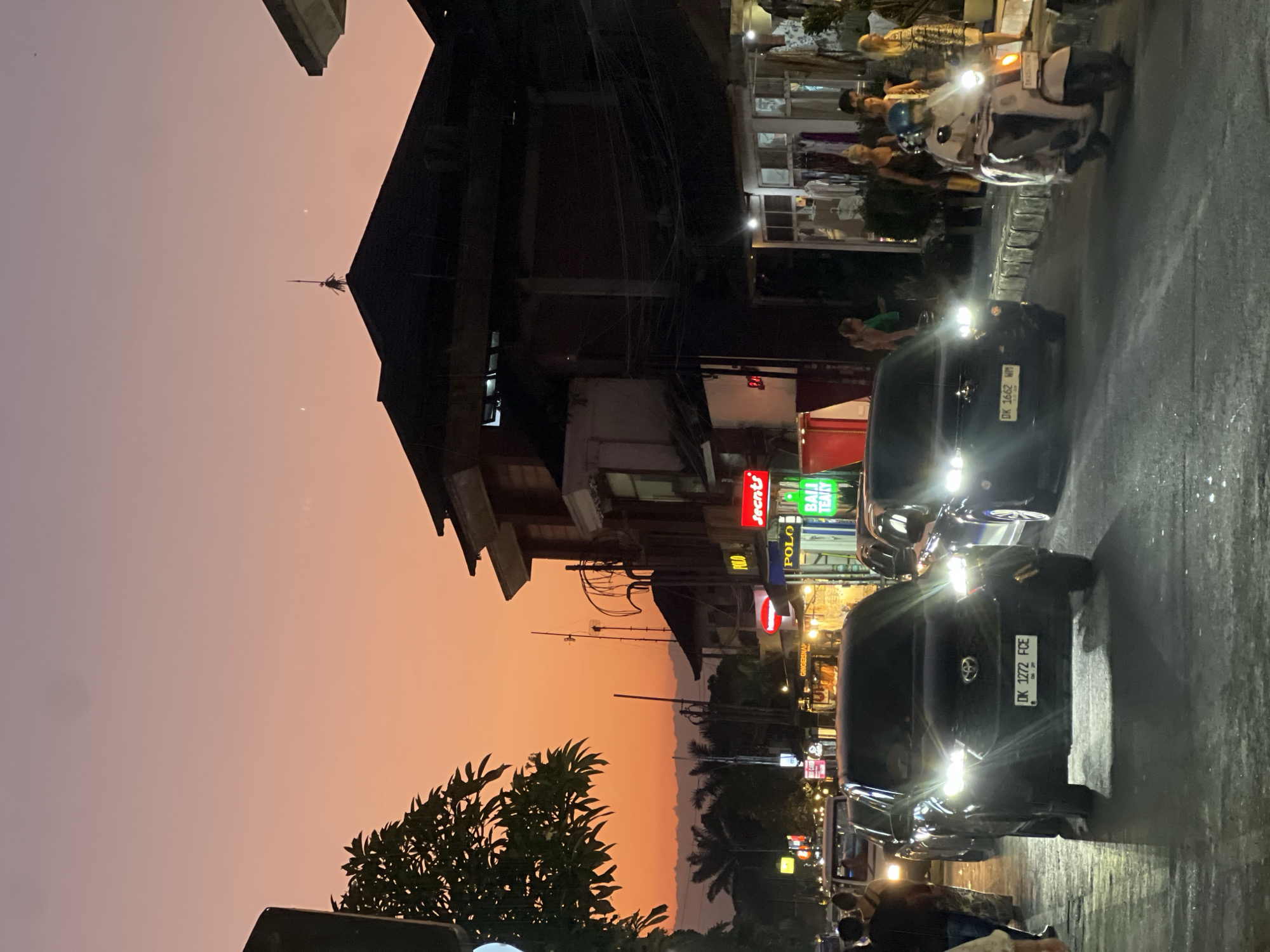 This image depicts a street scene in Ubud District, Bali, Indonesia, at twilight. The sky exhibits a gradient from pale pink-orange at the zenith to a deep orange at the horizon, where a distant mountain range is silhouetted. Two faint, crescent-shaped celestial bodies are visible in the upper sky above the right-side buildings.
On the left side of the frame, a dark, leafy tree partially obscures the upper corner. The right side features multi-story buildings, largely silhouetted, with one rooftop distinguished by a dark, spear-like finial or statue. At ground level, these buildings house shops, identifiable by illuminated signs such as "POLO," "Secrets," and "BALI TEA & COFFEE" (partially visible as "BALI TEATY"). Overhead utility wires crisscross above the street and buildings.
The paved road in the lower foreground appears wet or reflective. Two dark-colored vehicles with illuminated headlights are prominent, facing the viewer. The foremost vehicle is a black SUV/MPV with the Indonesian license plate "DK 1272 FCE." Behind it and to the right is another dark-colored vehicle displaying the license plate "DK 1662 WM." Further down the street, a white van is partially visible. On the right side of the road, a light-colored scooter with a rider wearing a helmet travels away from the viewer; its partially visible license plate begins "DK 5424 GM."
Several people populate the scene. On the left sidewalk, two individuals walk away from the viewer. On the right sidewalk, near the shops, three individuals are visible: two stand facing the shopfronts, and one in a patterned dress is further right. The scene is illuminated by the fading daylight and numerous artificial light sources from vehicle headlights, streetlights, and shop signs.