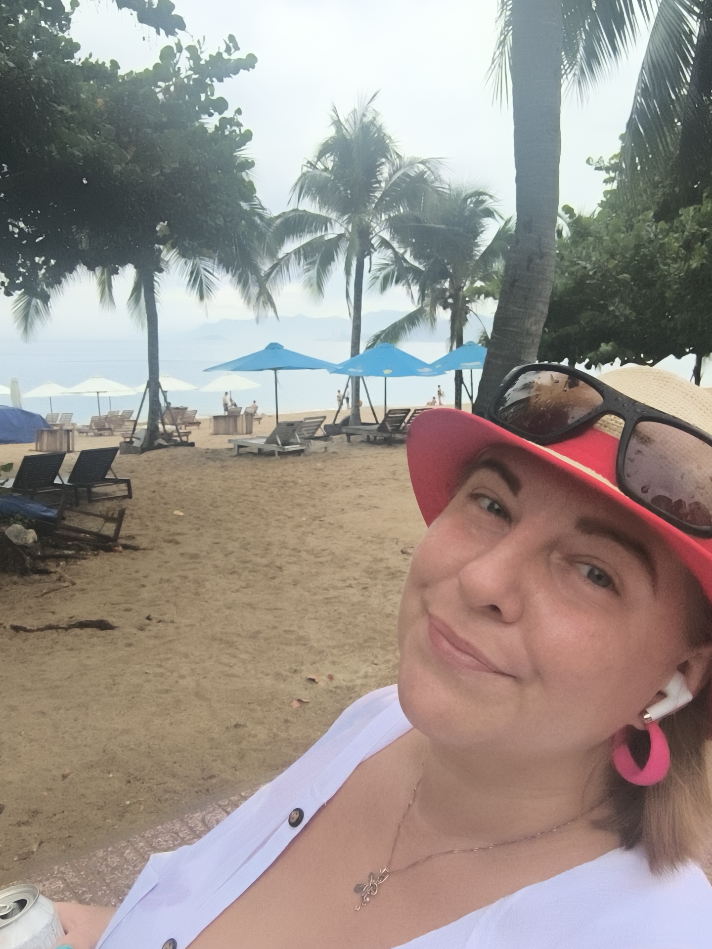 A woman wearing a red hat and sunglasses takes a selfie on a sandy beach in Nha Trang, Vietnam. She is wearing a white top and pink hoop earrings, and has white earbuds in her ears. In the background, palm trees, beach chairs, and umbrellas are visible along the coastline. The sky is overcast. A can of soda is held in her left hand.