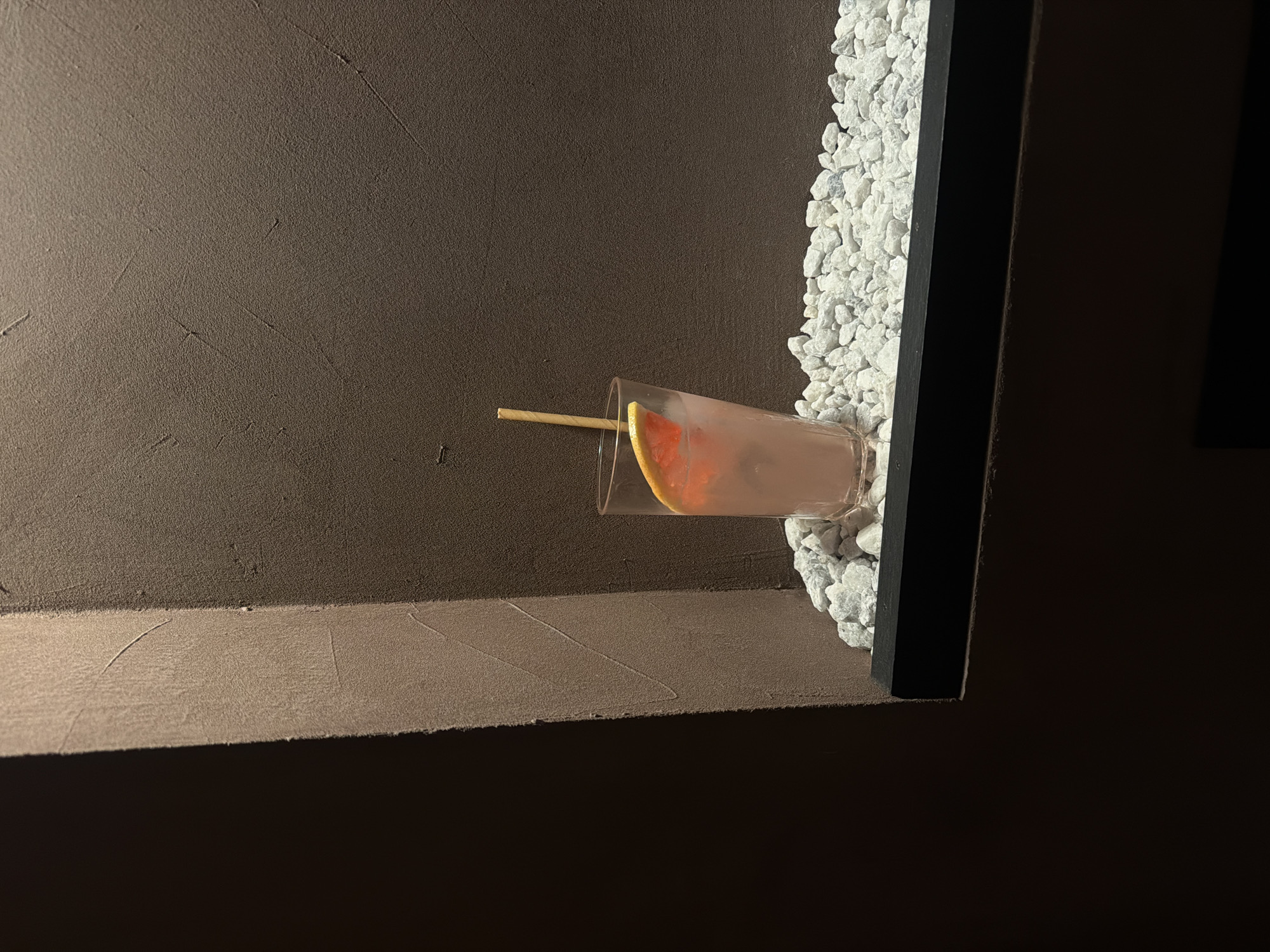 A clear glass, containing a light pink beverage with ice, a red grapefruit slice garnish, and a tan-colored straw, is centrally positioned on a dark, flat shelf. The shelf is covered with a layer of small, irregular white and light gray decorative pebbles. The background consists of two intersecting walls, exhibiting a textured, matte, dark brownish-gray finish, suggestive of stucco or plaster with subtle trowel marks. The wall immediately behind the glass is illuminated, highlighting its texture, while the wall to the left forms an architectural corner, with its surface facing the viewer appearing lighter due to illumination, contrasting with the deep shadow on its far left face. The scene is dimly lit, focusing attention on the drink and the textural details of the surrounding surfaces. No individuals or active interactions are depicted. This scene is located in the city of Huddinge, Sweden.