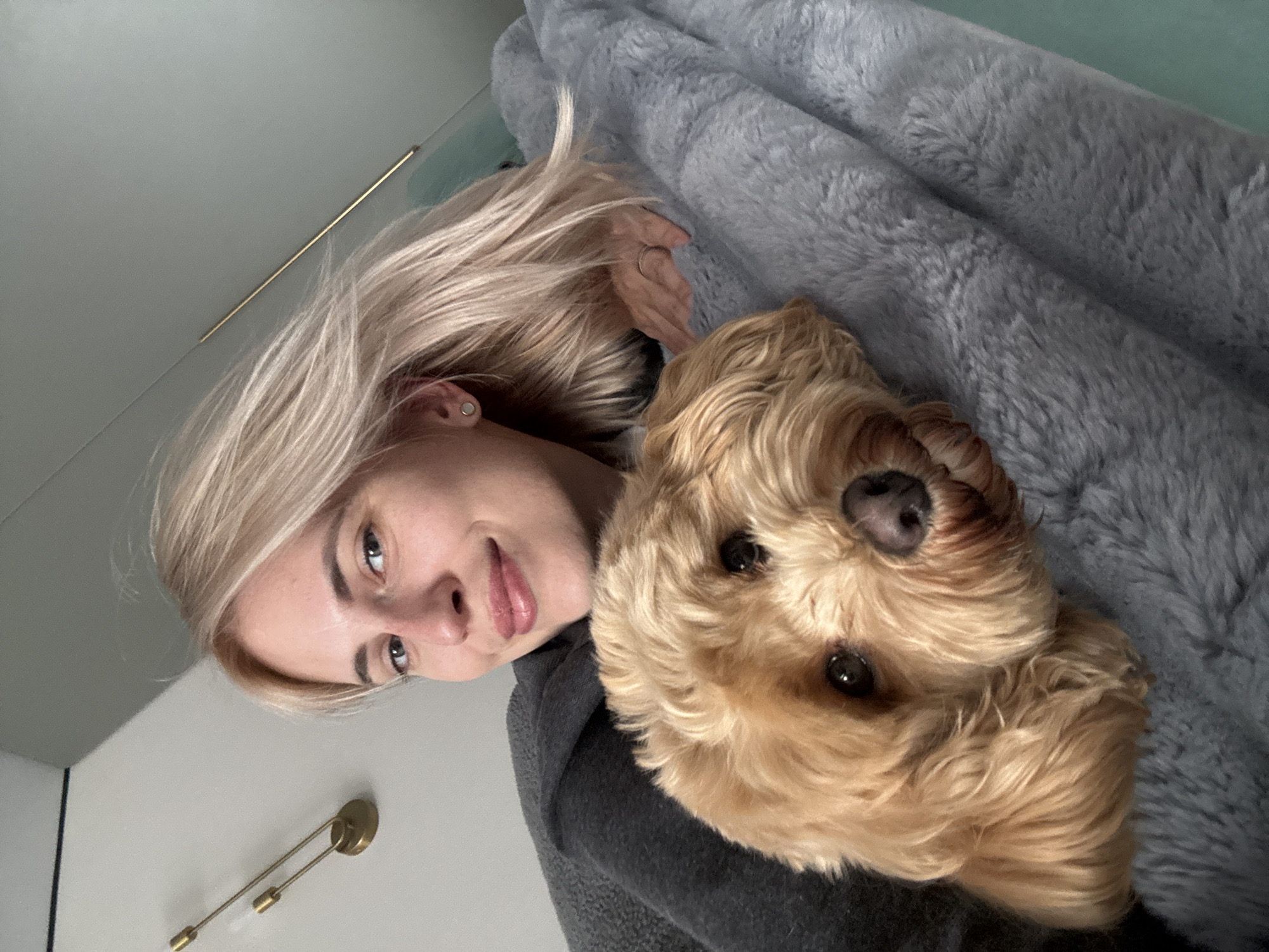 A woman with short blonde hair is lying down, next to a golden-colored dog, on a gray fuzzy blanket. The woman is looking at the camera with a slight smile. She is wearing a dark gray sweater. The dog is also looking at the camera. A wall with a gold-colored light fixture is visible in the background. The location is unspecified, and there is no information that can be used to determine this image was taken in Saratov, Russia.