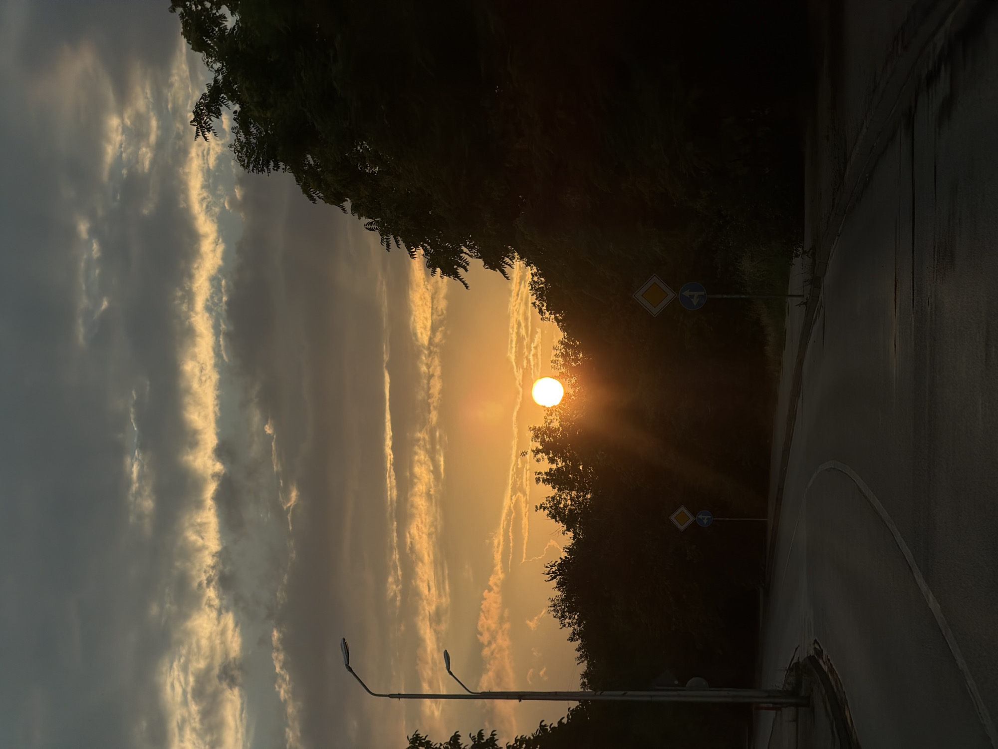 The image captures a sunset over a curved road, likely in the city of Lovech, Bulgaria. The sun, a bright orb, is partially obscured by silhouetted trees on the right and a band of clouds across the sky. The sky transitions from a dark, cloudy grey at the top to a vibrant orange and yellow hue near the horizon, with rays of sunlight emanating from the sun. On the left side of the road, two streetlights stand tall, their lamps illuminated. The road itself appears to be asphalt, with a white line marking its edge. Two road signs are visible: one diamond-shaped yellow sign above a circular blue sign with arrows indicating directions, placed on the side of the road. The surrounding area is densely covered in dark trees. The overall scene conveys a sense of twilight and the end of the day.