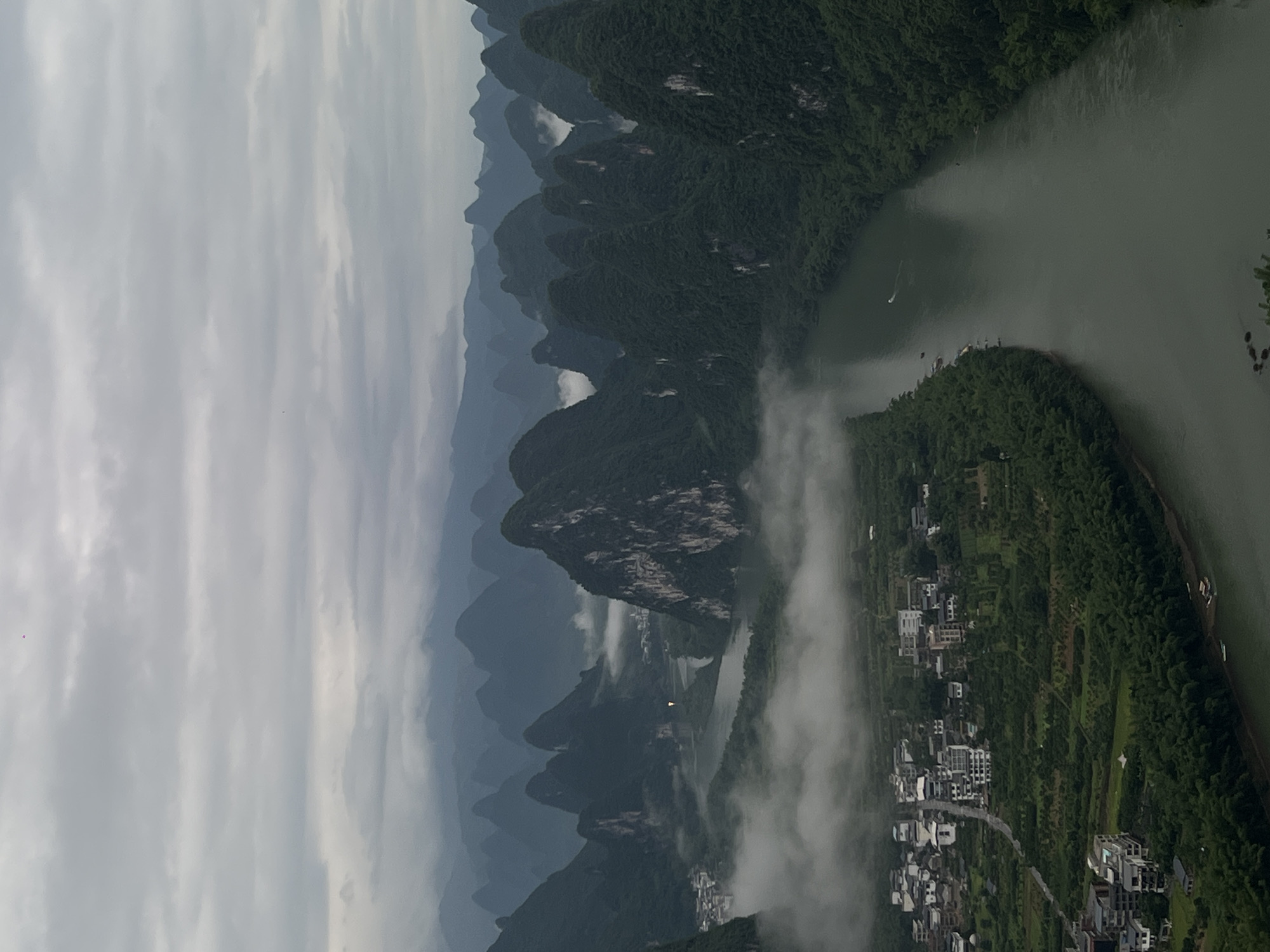 The image depicts a landscape in Guilin, China. Karst mountains rise in the background, partially obscured by mist. A river winds through the landscape, with a small boat visible on the water. A town with buildings is situated along the riverbank, amidst lush greenery. The sky is overcast with grey clouds.