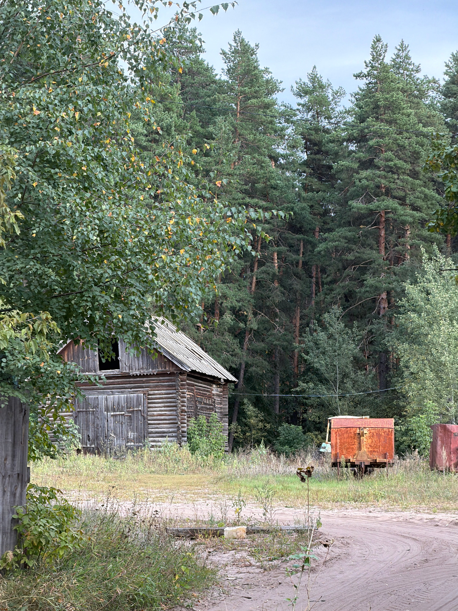 In the village of Uzukovo, Russia, a weathered wooden log cabin with a corrugated metal roof stands to the left of a dirt road. The cabin has large double doors and an open, dark window on its upper story. The surrounding area is overgrown with tall grass and weeds. In the background, a dense forest of pine trees dominates the landscape. To the right of the cabin, on the other side of the road, is a rusty, orange metal container on wheels, and further to the right, a portion of a red metal silo is visible. The sky is overcast with a light blue hue.