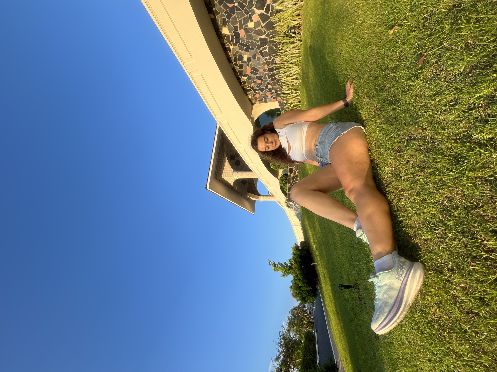 A woman with curly brown hair sits on a grassy slope in front of a building with a mosaic wall under a clear blue sky. She is wearing a white crop top and denim shorts, with light blue and purple athletic shoes. Her legs are crossed, and her right arm rests on the grass behind her. The building appears to be modern with ornate architectural details and a long, overhanging roof. The grass is neatly trimmed and illuminated by the warm light of what appears to be a sunset. In the background, trees and palm trees are visible along a paved road. The location is specified as Phu Thuy Ward, Vietnam.