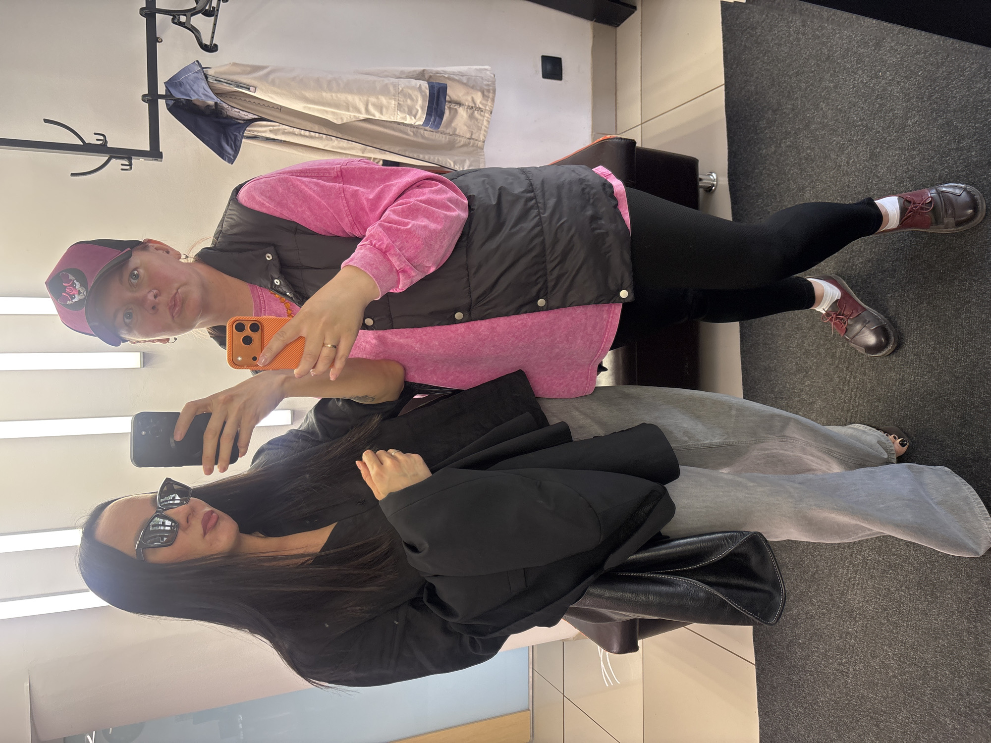 Two women are taking a selfie in a room that appears to be a dressing room or a retail space. The woman in the foreground is wearing sunglasses, a black jacket, light grey jeans and sitting with a black leather bag. The woman in the background is holding an orange phone, wearing a black puffer vest, a pink shirt, black leggings, red shoes, a maroon and black hat, and standing. Clothes hang in the background. The location is Chaikovsky, Russia.